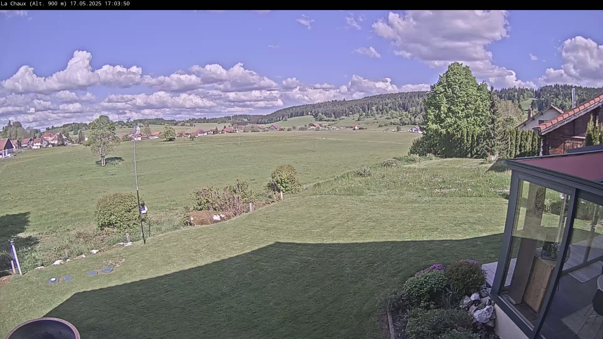 A partly cloudy sky overlooks a sprawling green field, a small village in the distance, and a portion of a house with a glass enclosure in the foreground.