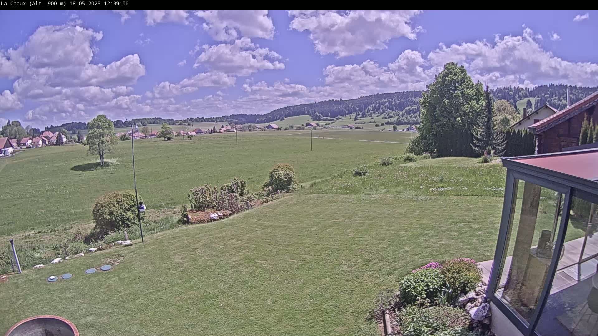 A partly sunny day reveals a sprawling green field, a small village in the distance, and a garden with a glass enclosure in the foreground.