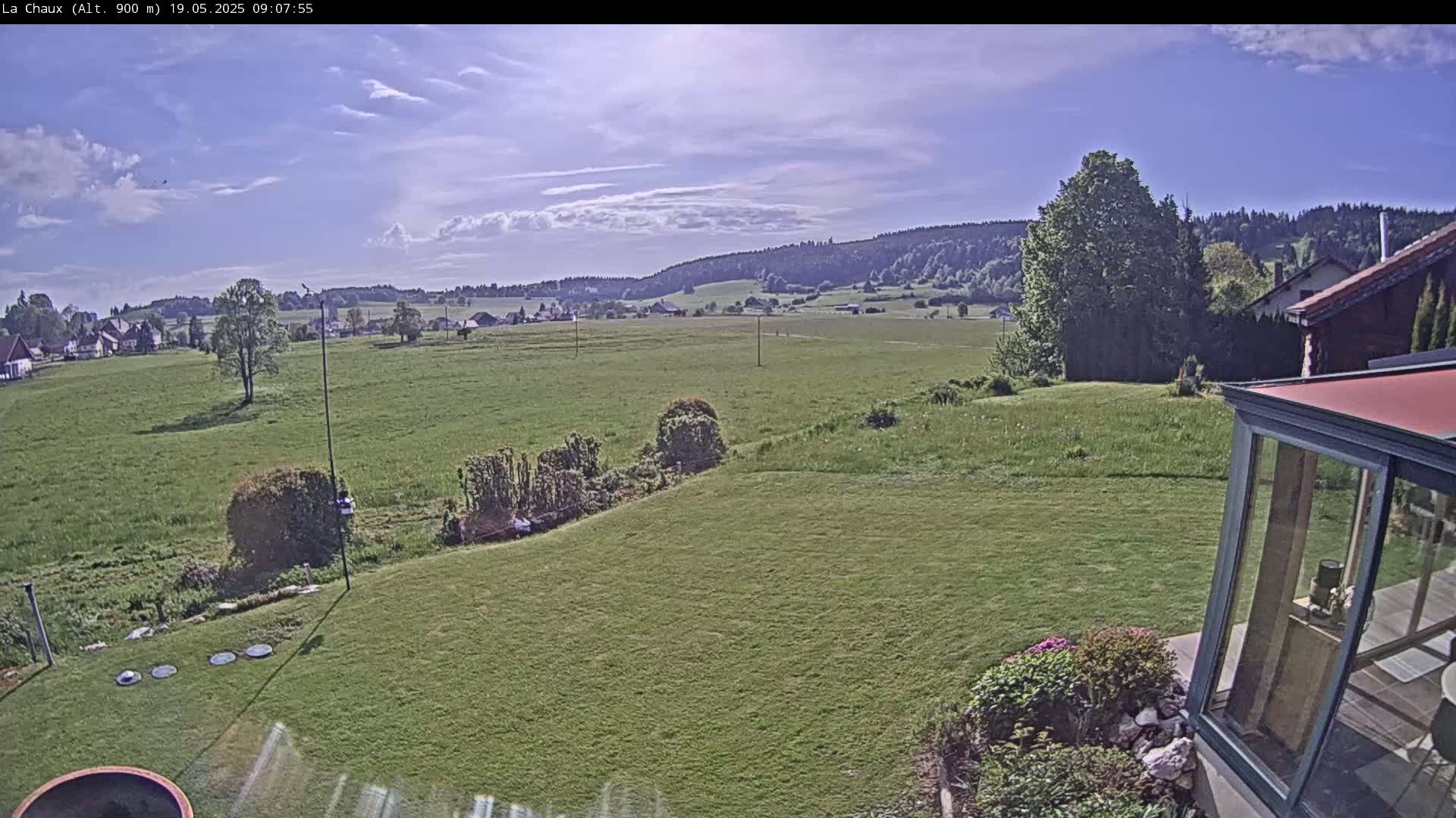 A partly cloudy day reveals a view of a green field extending to a line of trees and distant hills, seen from a backyard with a modern glass structure and manicured lawn.