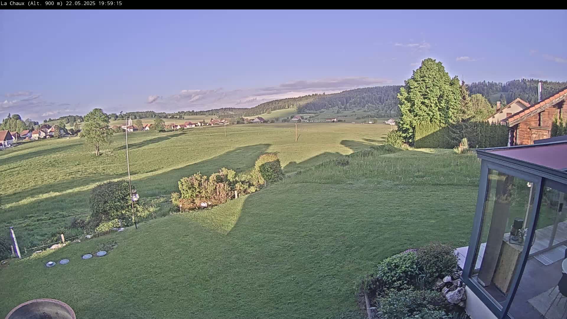 A verdant lawn slopes down to a field, beyond which lies a village nestled at the foot of a forested hill under a mostly clear, light blue sky.