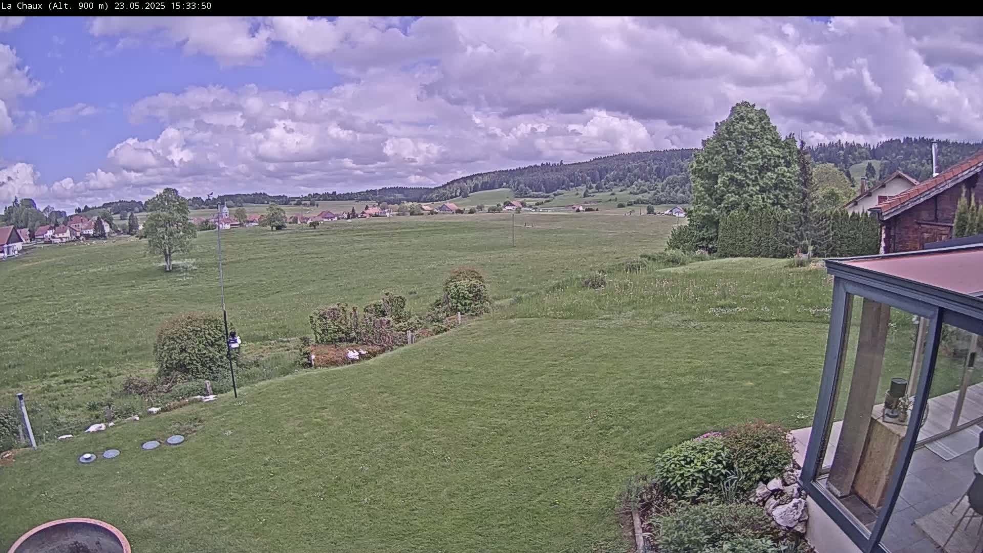 A partly cloudy day reveals a sprawling green field, a small village in the distance, and a garden with a modern glass structure nearby.