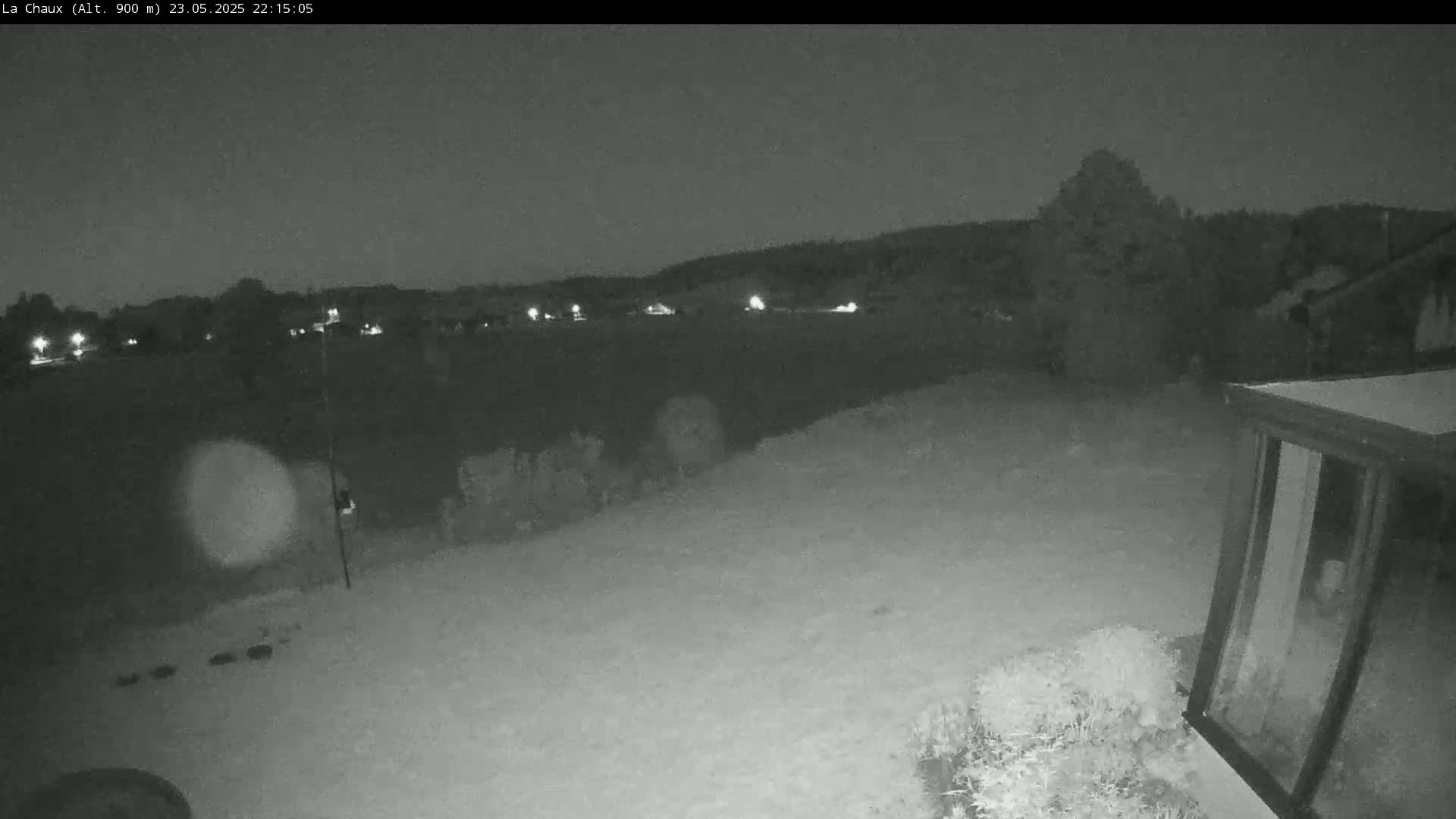 A nighttime, low-light view of a backyard shows a snow-covered ground, some shrubs, a distant line of houses with lights, and a glass enclosure.