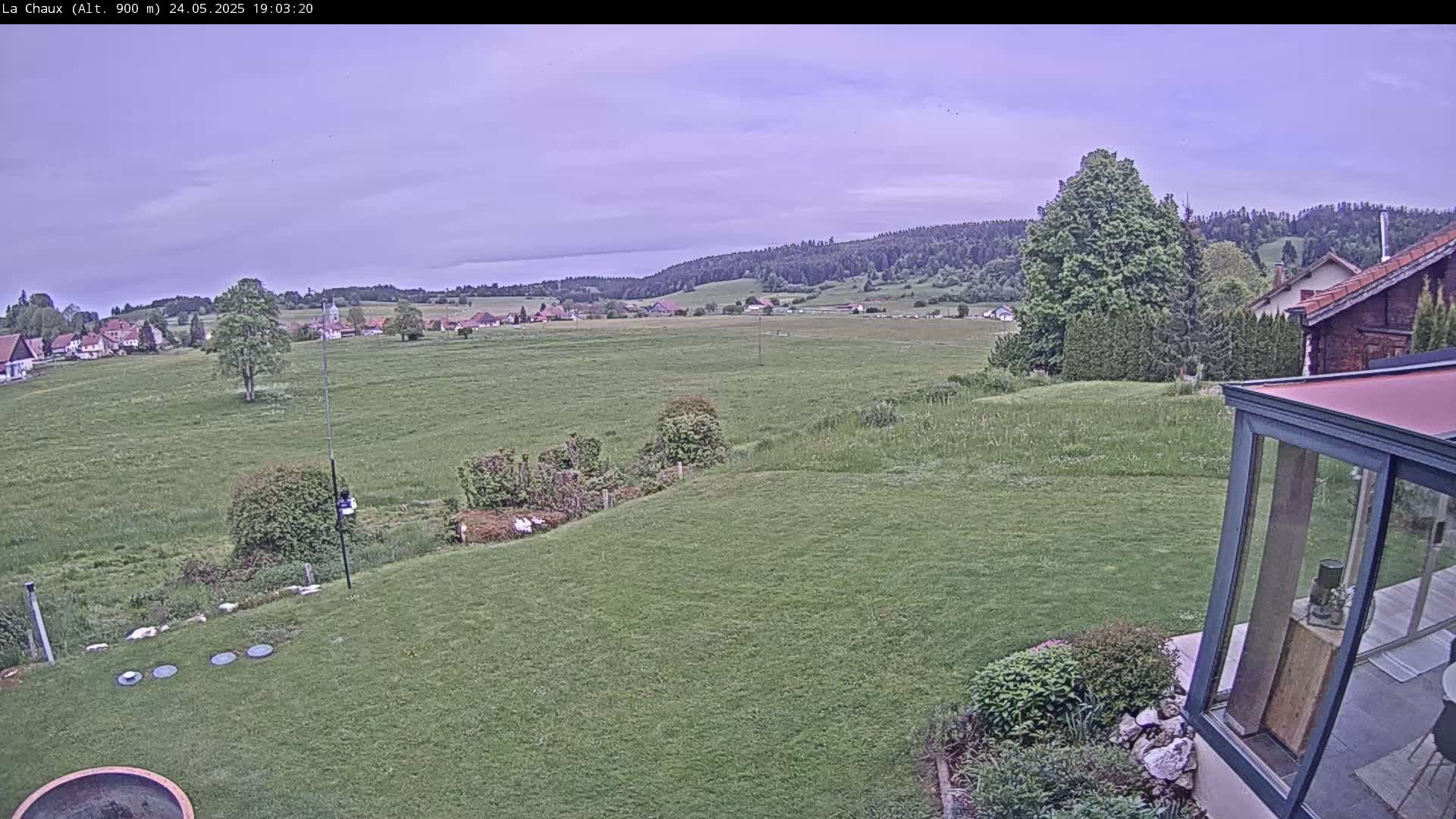 A mostly cloudy view of a green field and distant village, seen from a backyard with a glass sunroom and several round objects on the lawn.
