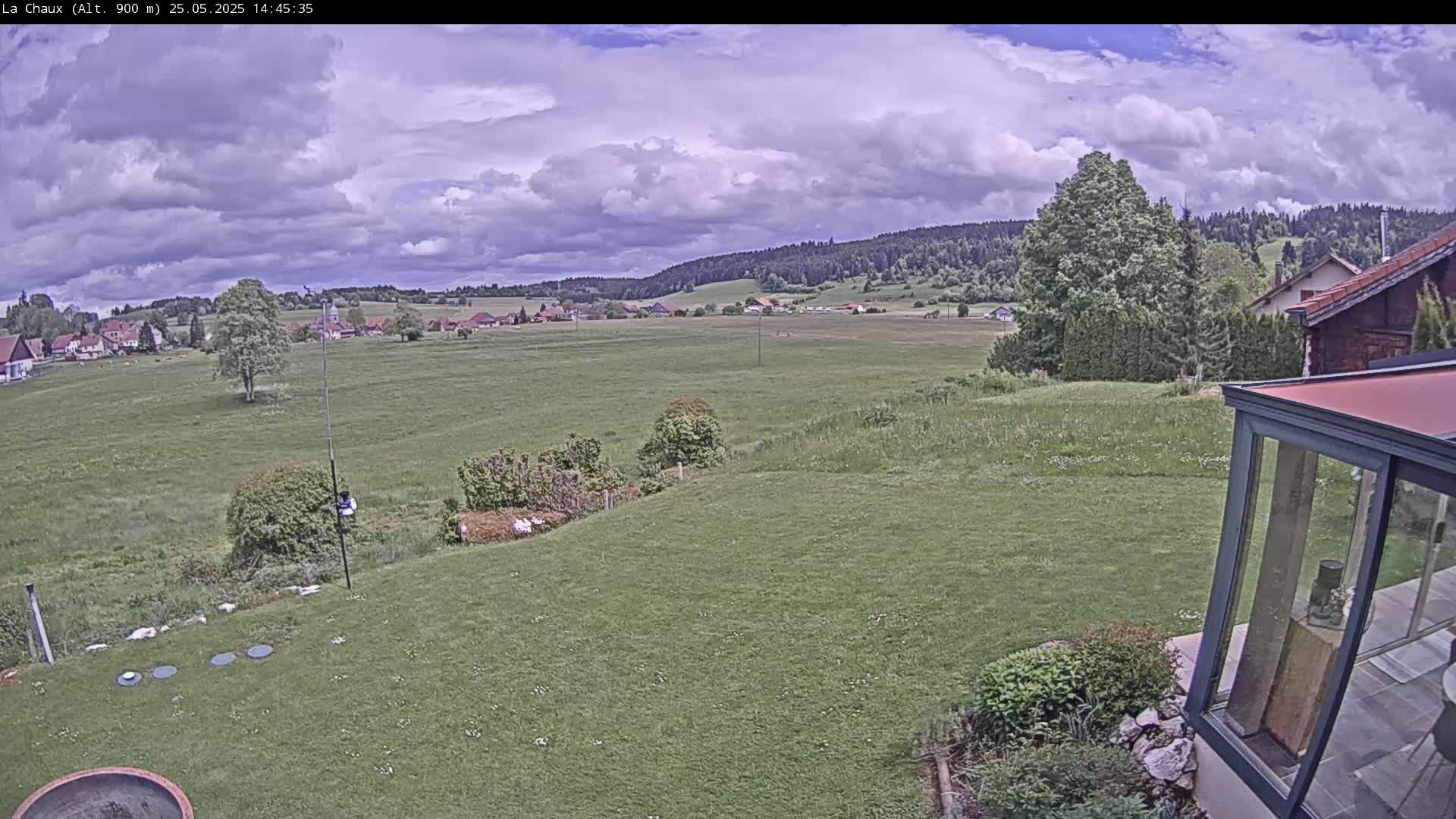 A sprawling green field extends to a village nestled among rolling hills under a partly cloudy sky.