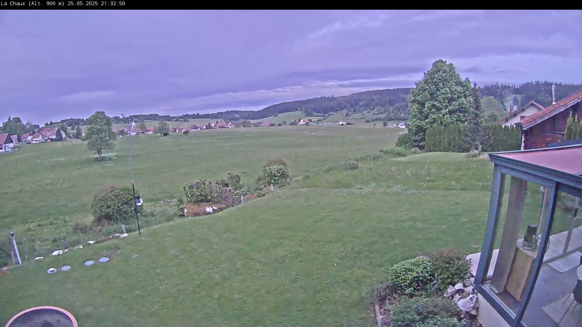 A grassy yard slopes down to a wide, flat field, beyond which lies a village nestled amongst hills under a dusky, cloudy sky.