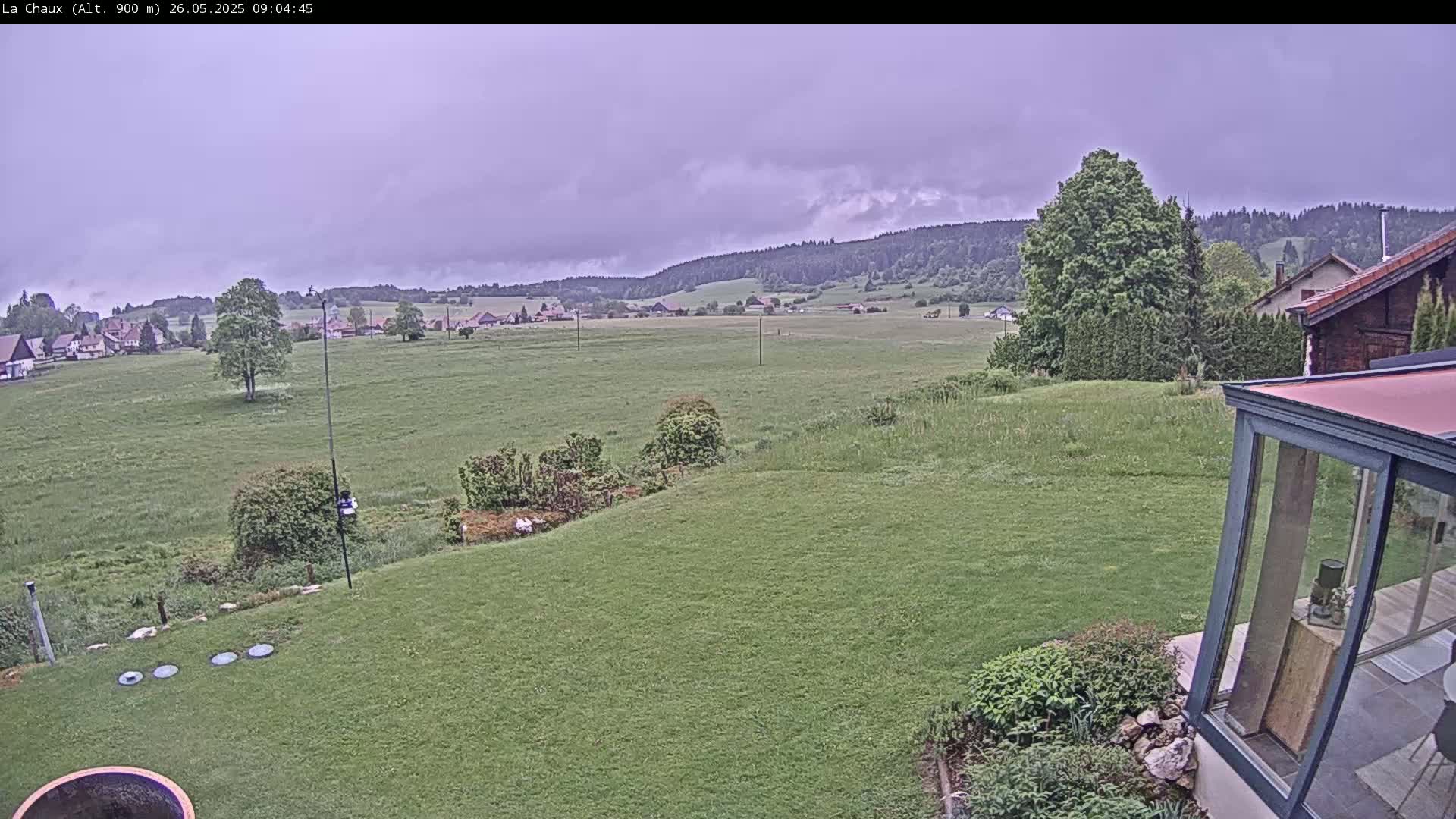 A green lawn slopes down to a flat expanse of fields and a village nestled against a hillside under a cloudy sky.