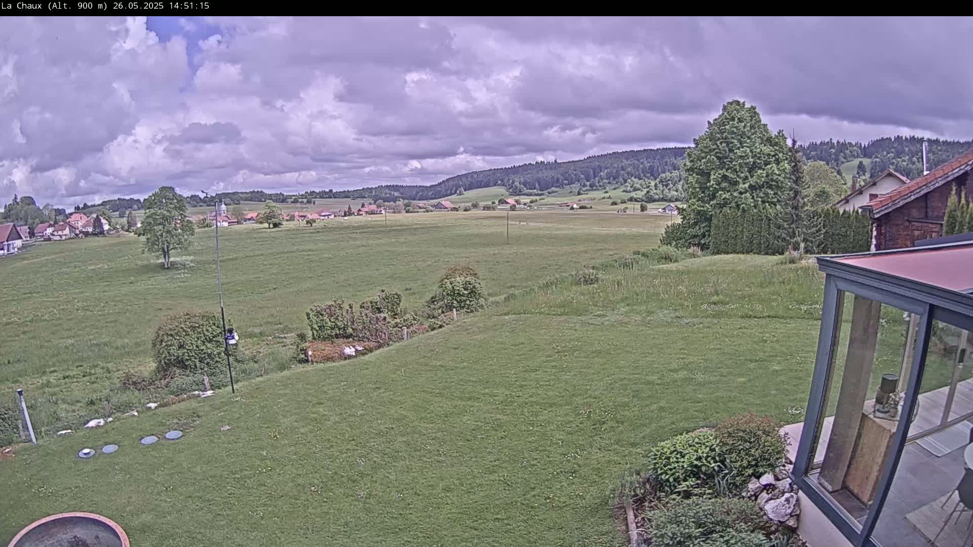 A sprawling green lawn with a distant village and forest under a partly cloudy sky.