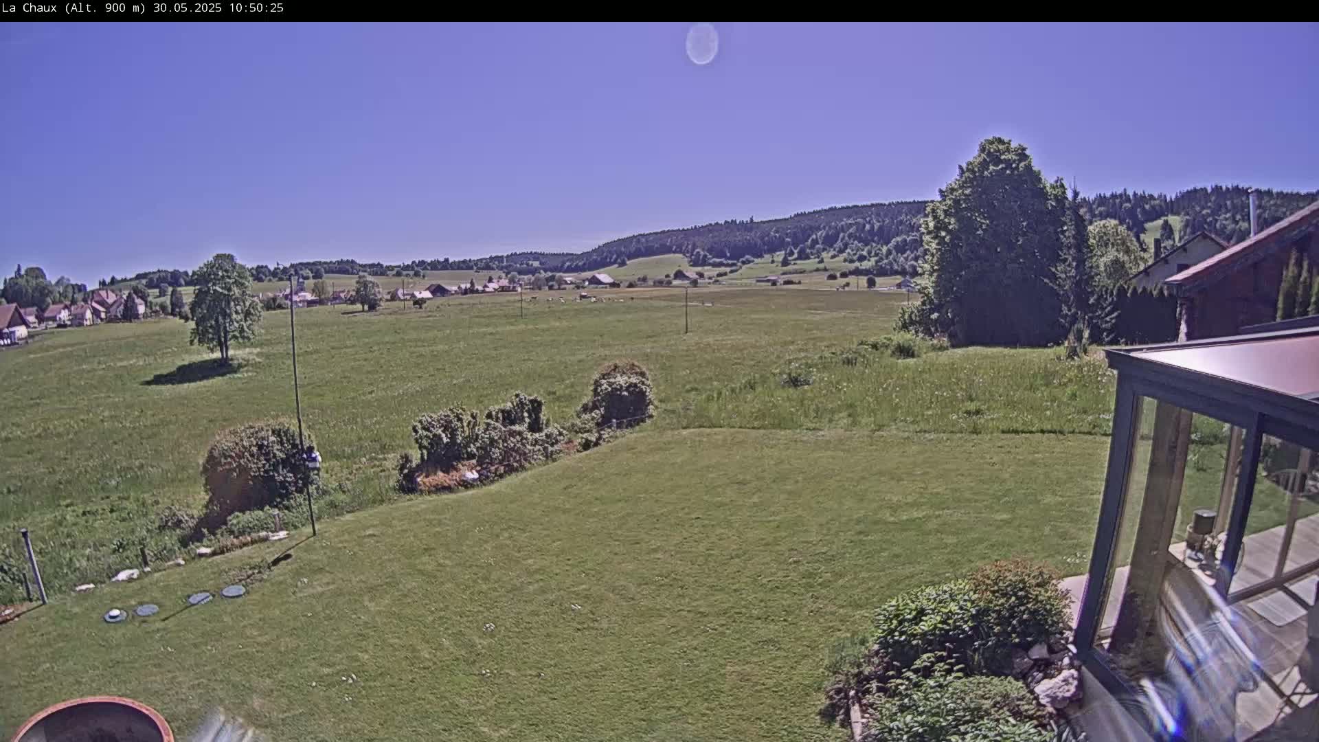 A grassy yard slopes down to a field, beyond which a village sits nestled in front of a line of forested hills under a clear blue sky.