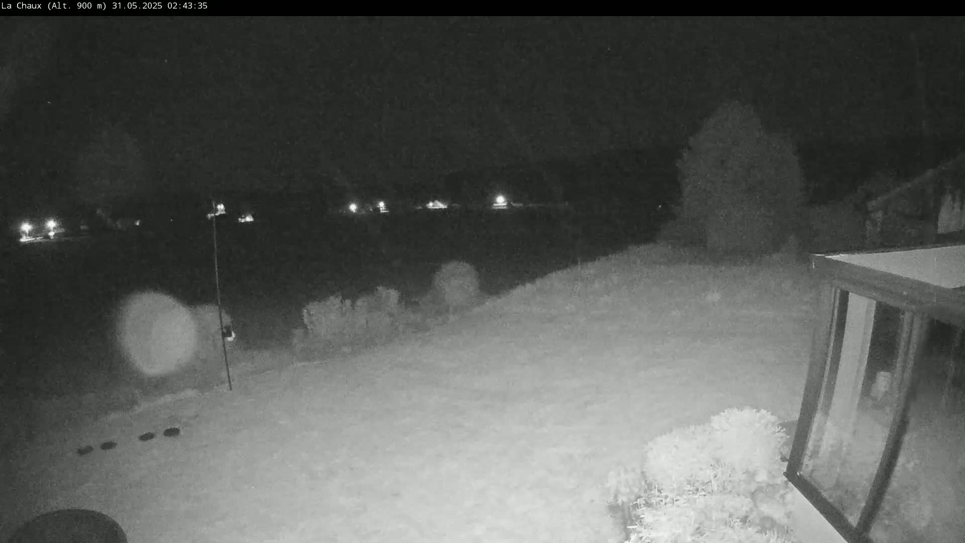 A snow-covered backyard at night is illuminated by distant lights, with a tall pole and part of a building visible in the foreground.