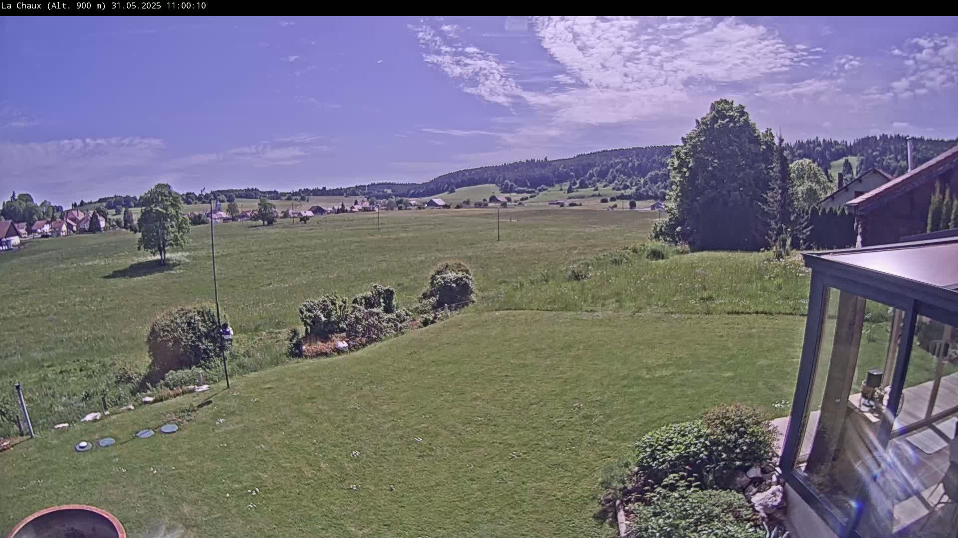 A sunny day reveals a verdant landscape featuring a sprawling green field, a distant village nestled among rolling hills, and a portion of a backyard with a modern glass enclosure.