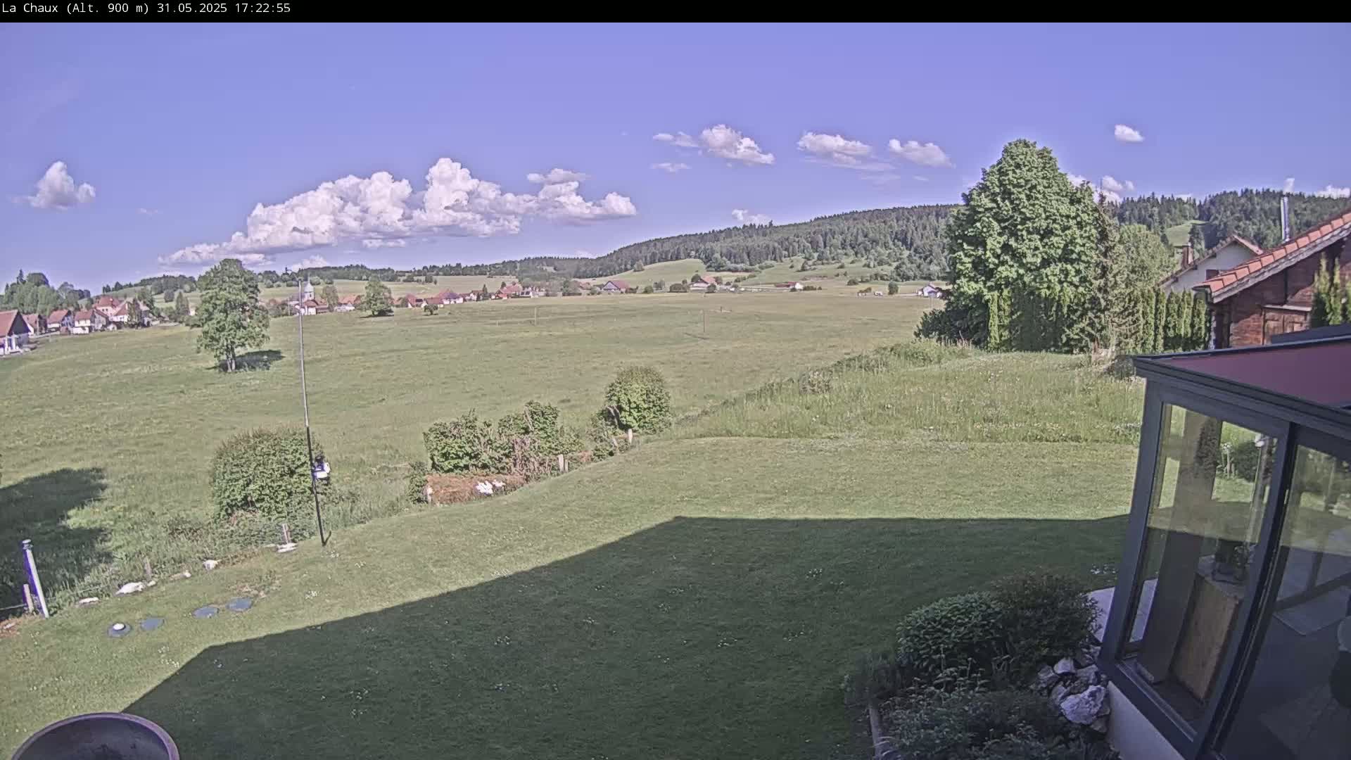 A partly sunny day reveals a sprawling green field, a distant village nestled in a valley, and a portion of a residential property with a sunroom, all backed by a dark forest.