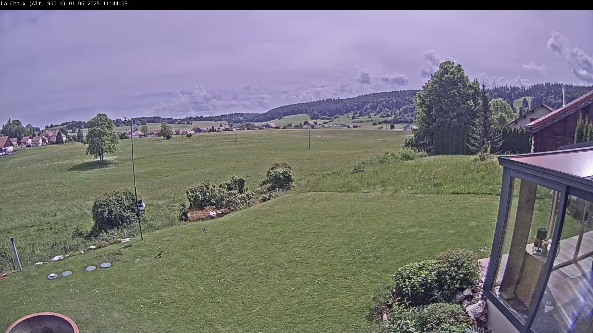 A partly cloudy sky overlooks a verdant expanse of fields and a village nestled in a valley, bordered by dark green hills, with a modern glass conservatory and lawn in the foreground.