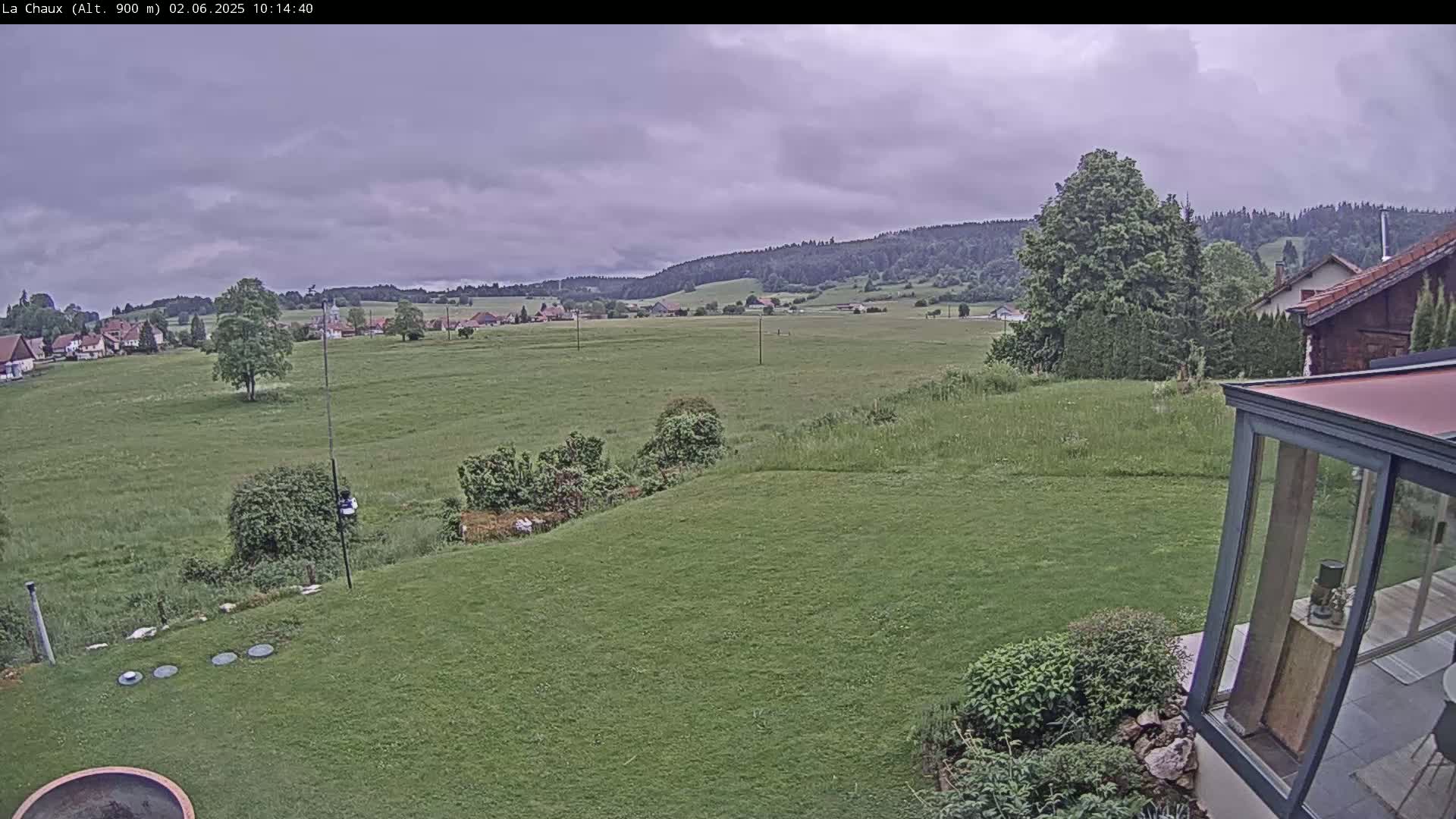 A grassy yard slopes down to a field and distant village under an overcast sky.
