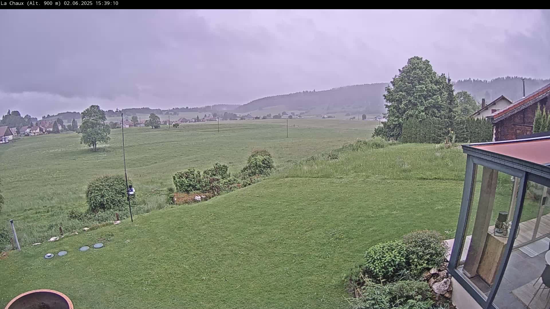 A grassy yard slopes down to a field under an overcast sky, with a village and hills in the distance.