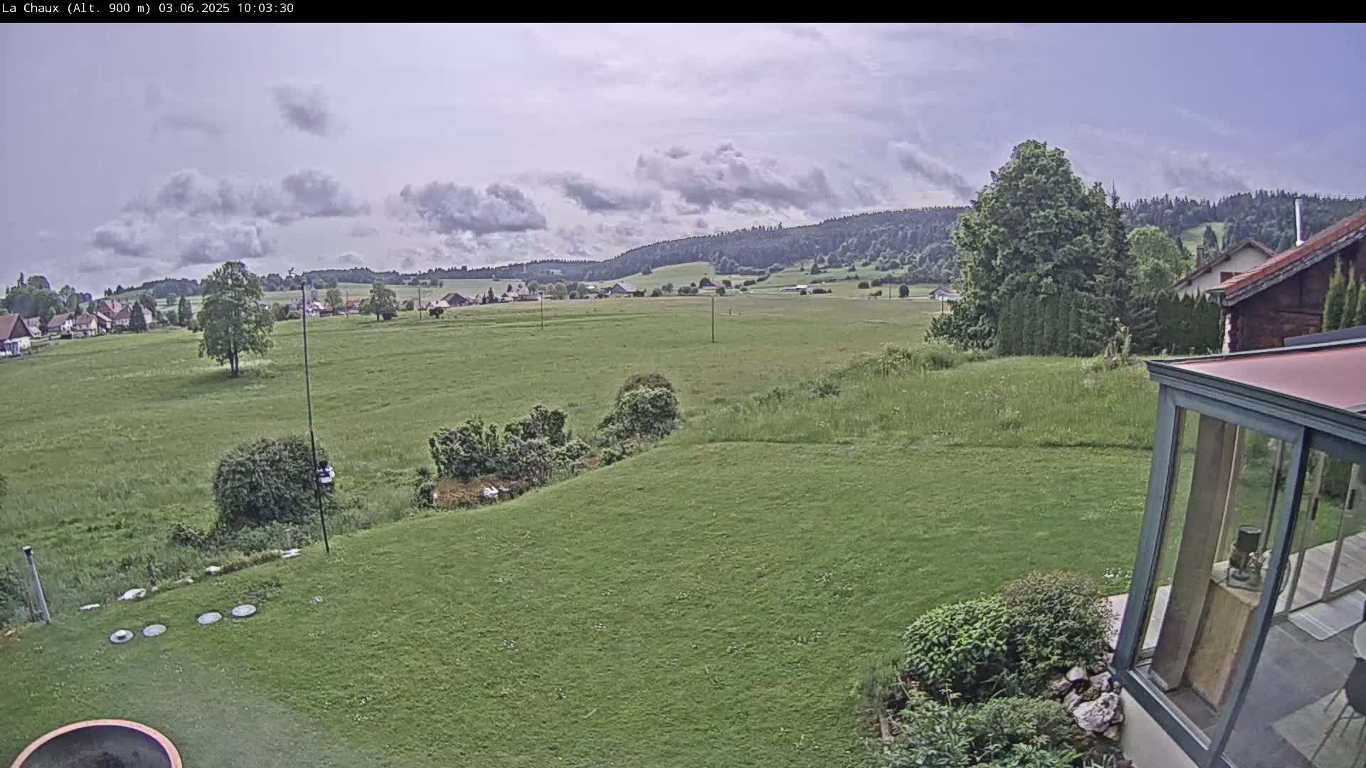 A partly cloudy day reveals a verdant landscape of rolling hills, a sprawling green field, and a portion of a house with a glass sunroom, all observed from a backyard vantage point.