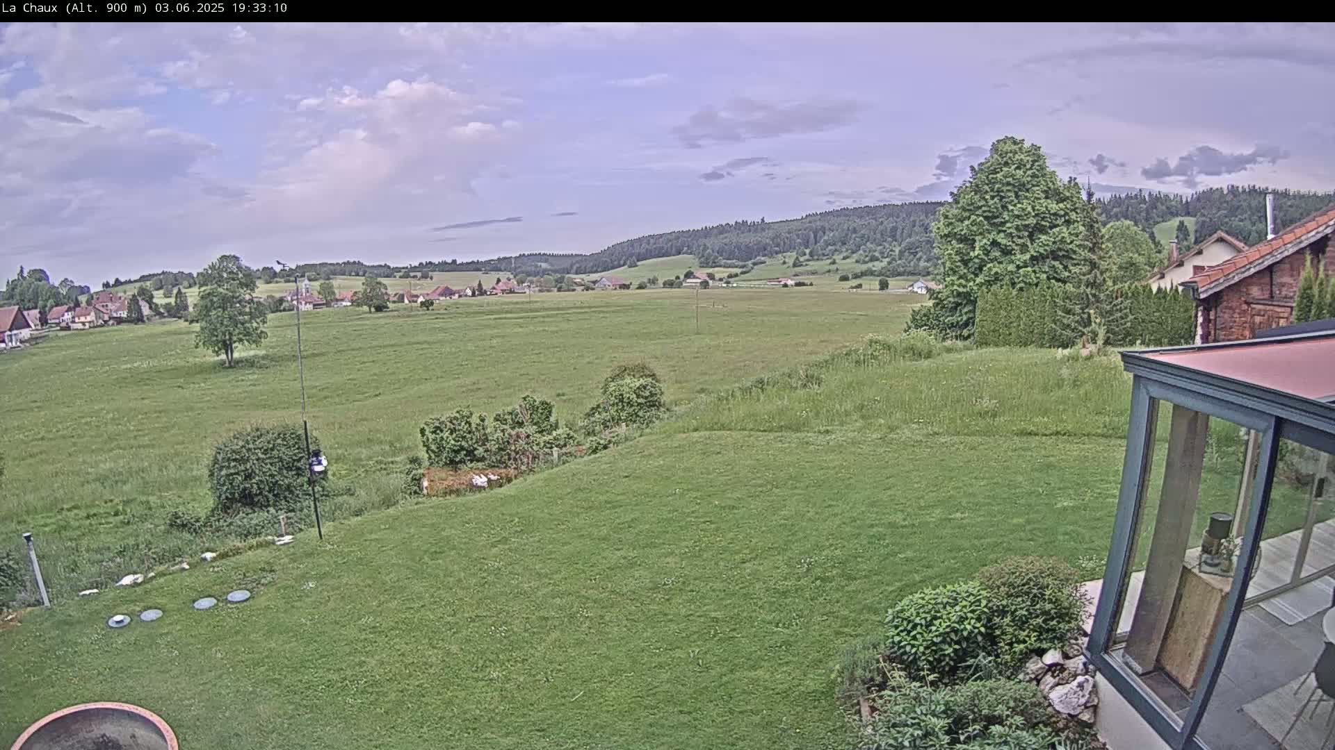 A green grassy yard slopes down to a flat field, beyond which lies a small village nestled against a line of dark green hills under a partly cloudy sky.