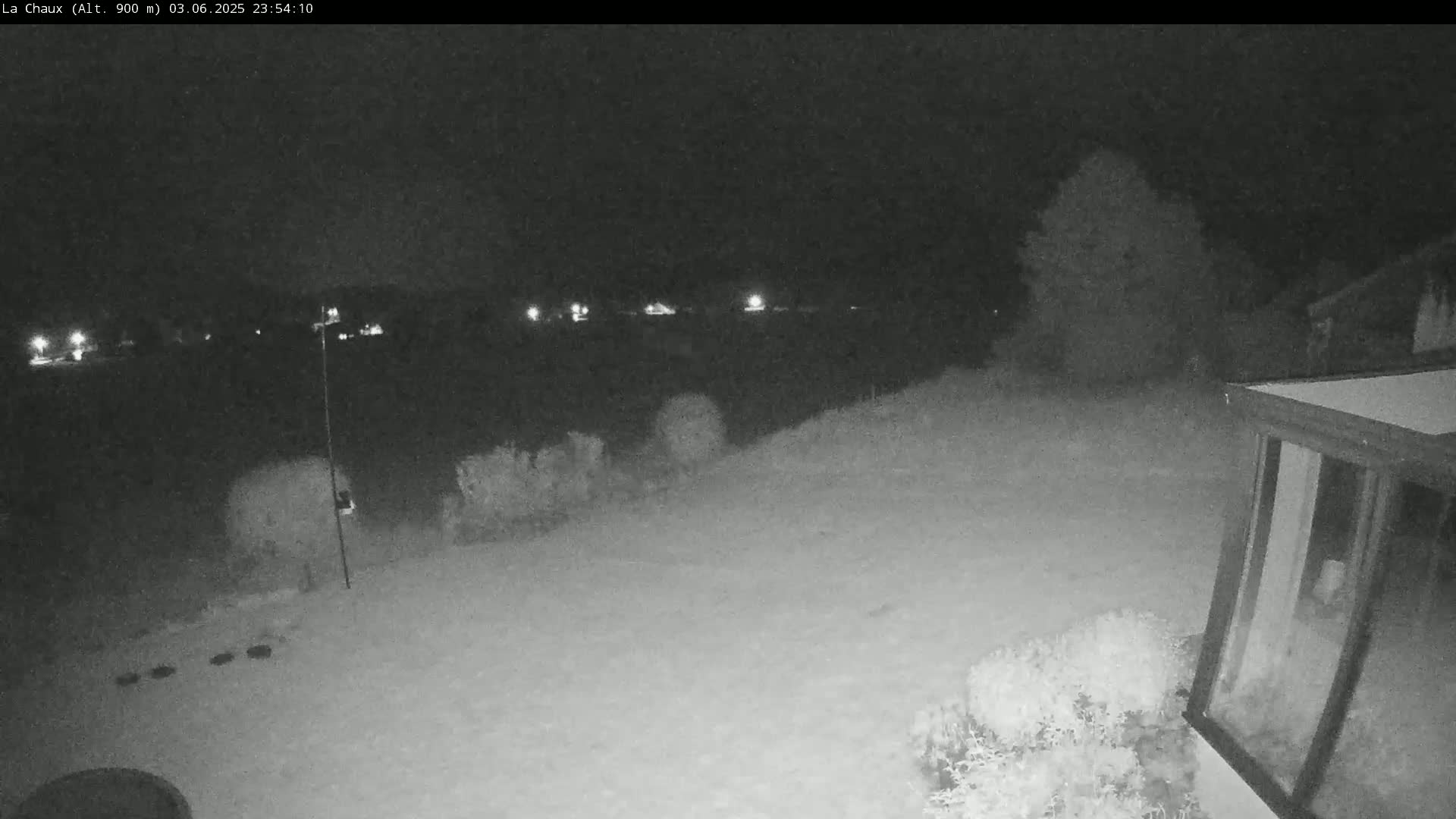 A snow-covered backyard at night is viewed from a security camera, showing a house and distant lights.