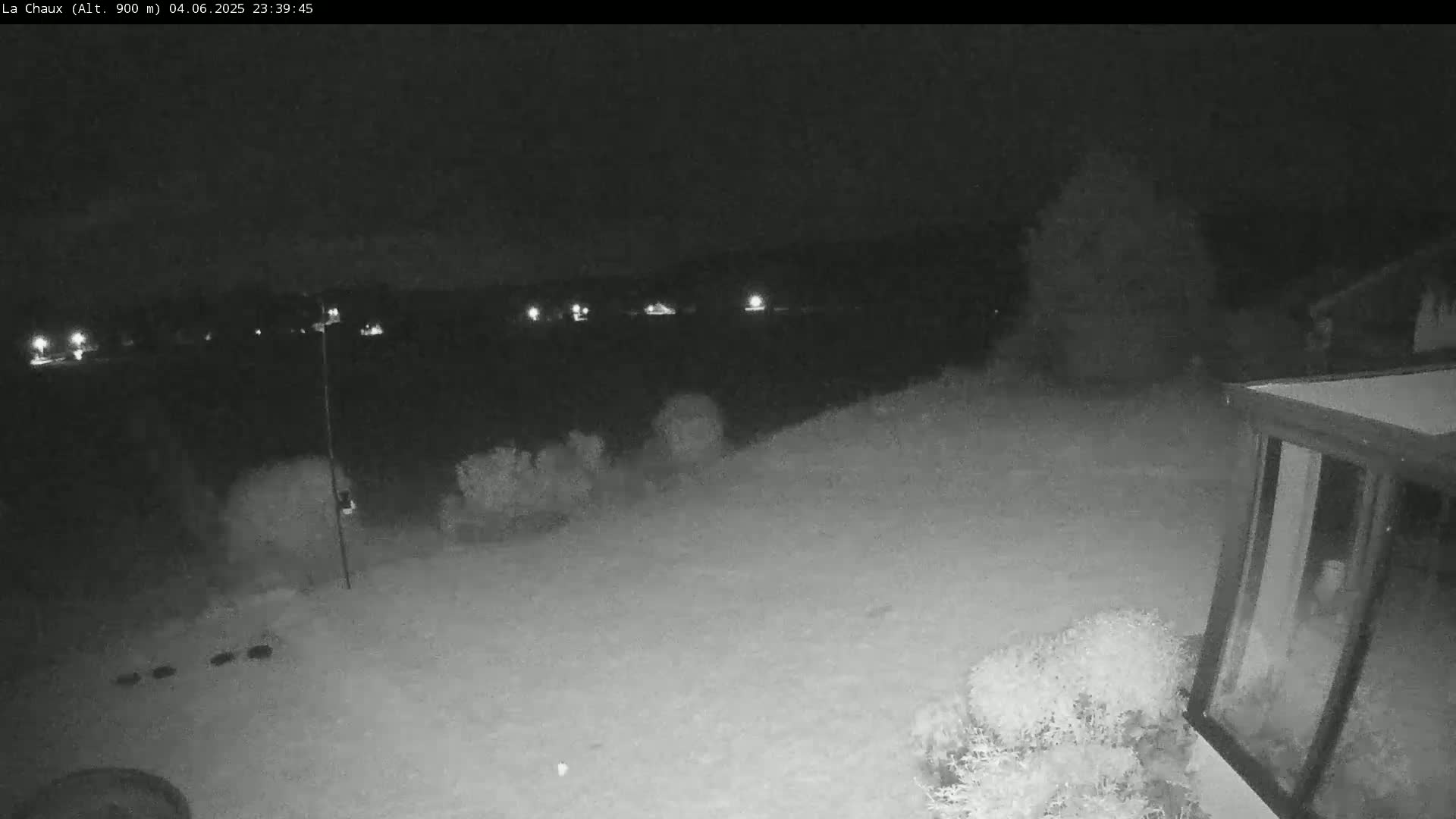 A snow-covered backyard at night is viewed from a security camera, showing a corner of a house and distant lights.