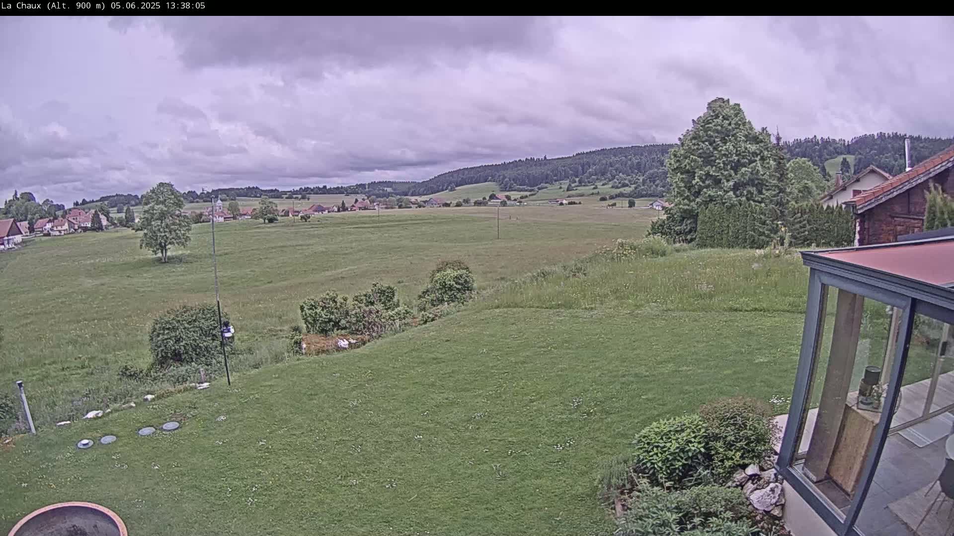 A grassy yard with a distant village and rolling hills under a cloudy sky is seen from a backyard perspective.