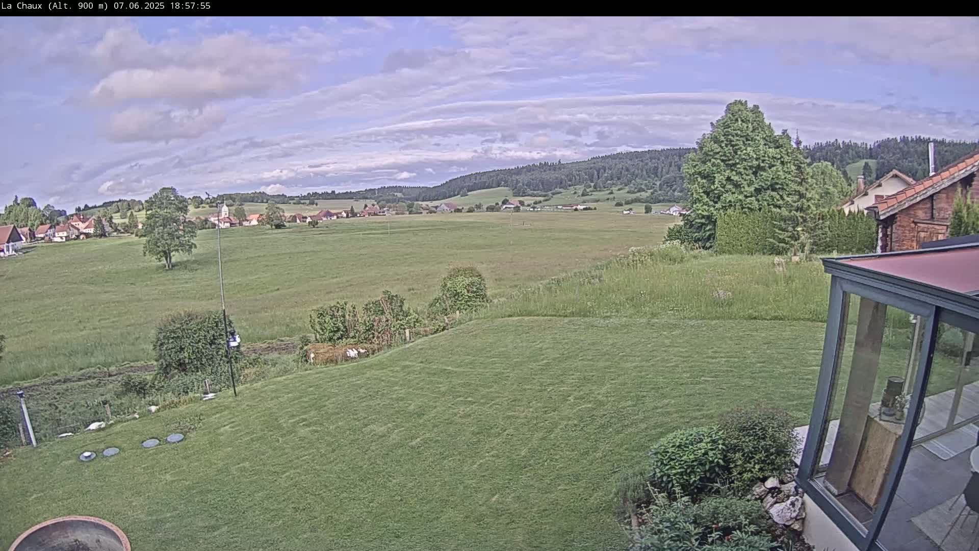 A sprawling green lawn slopes down to a verdant field and distant village under a partly cloudy sky.