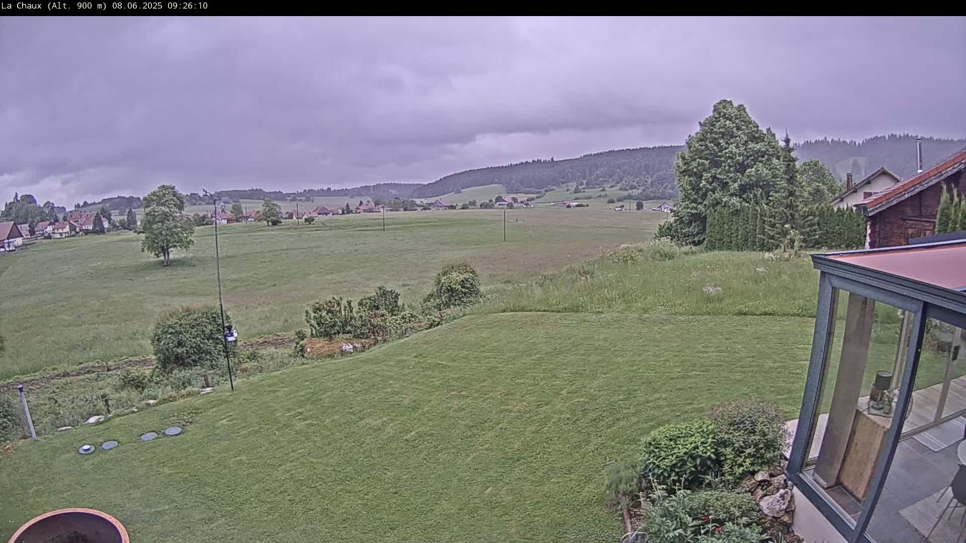 A grassy backyard overlooks a field and distant village under an overcast sky.