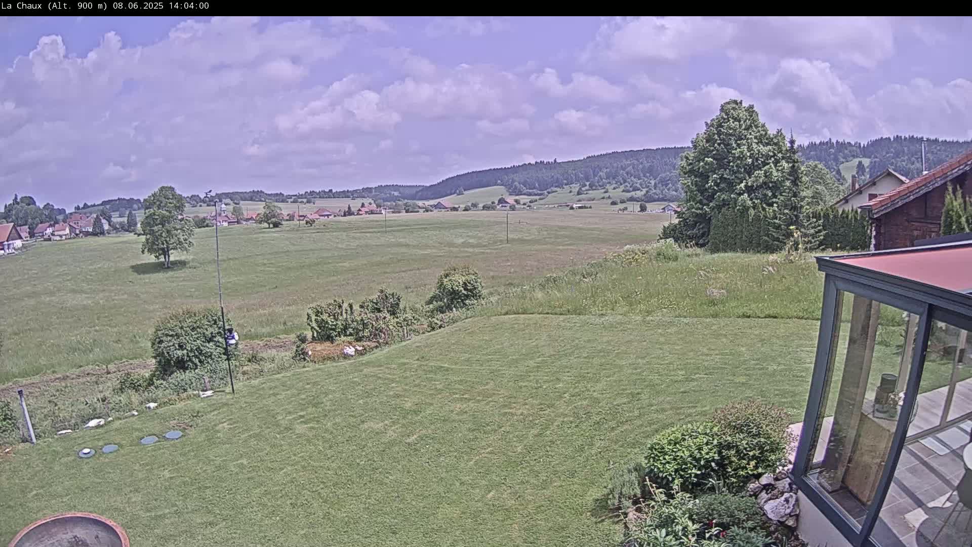 A partly cloudy day reveals a sprawling green lawn, a distant village nestled amongst fields, and a forested hillside under a mostly sunny sky.