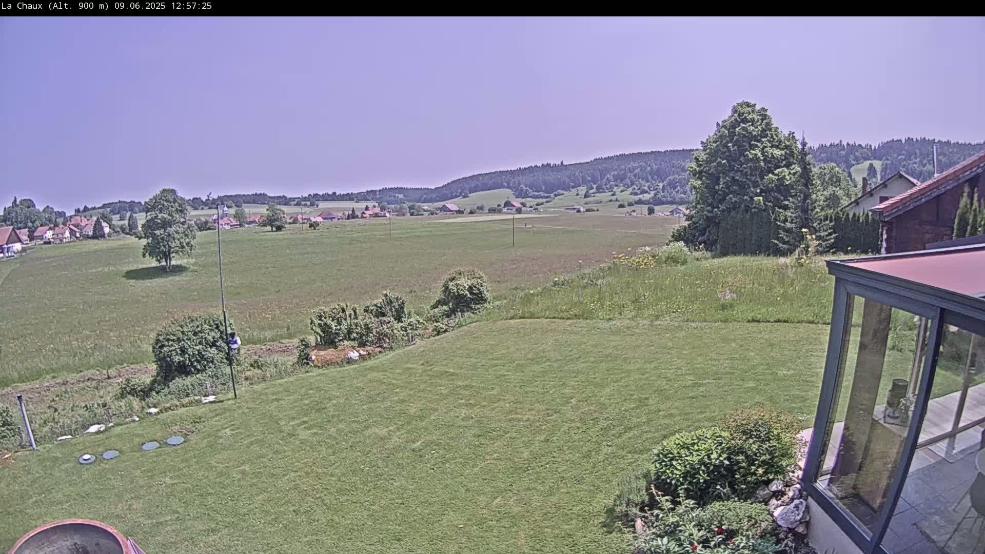 A grassy backyard scene under a clear sky overlooks a sprawling green field, a small village, and distant forested hills.