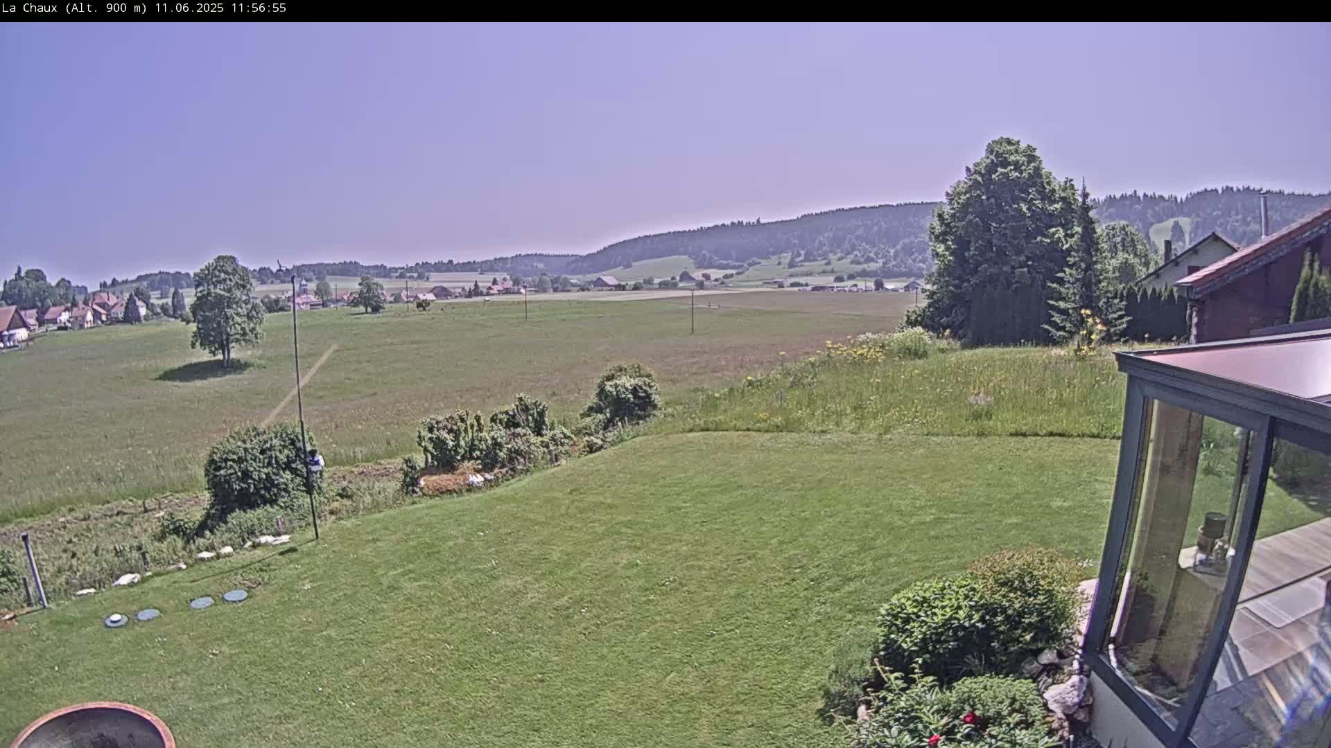 A grassy yard slopes down to a field, beyond which lies a village nestled against a line of forested hills under a clear blue sky.