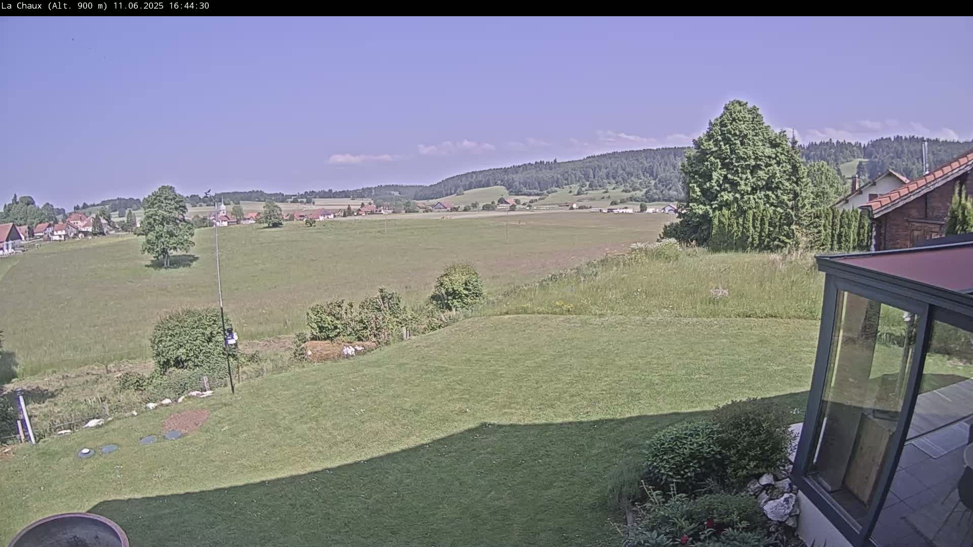 A sunny view of a grassy backyard, overlooking a village nestled in a valley surrounded by rolling hills and forests.