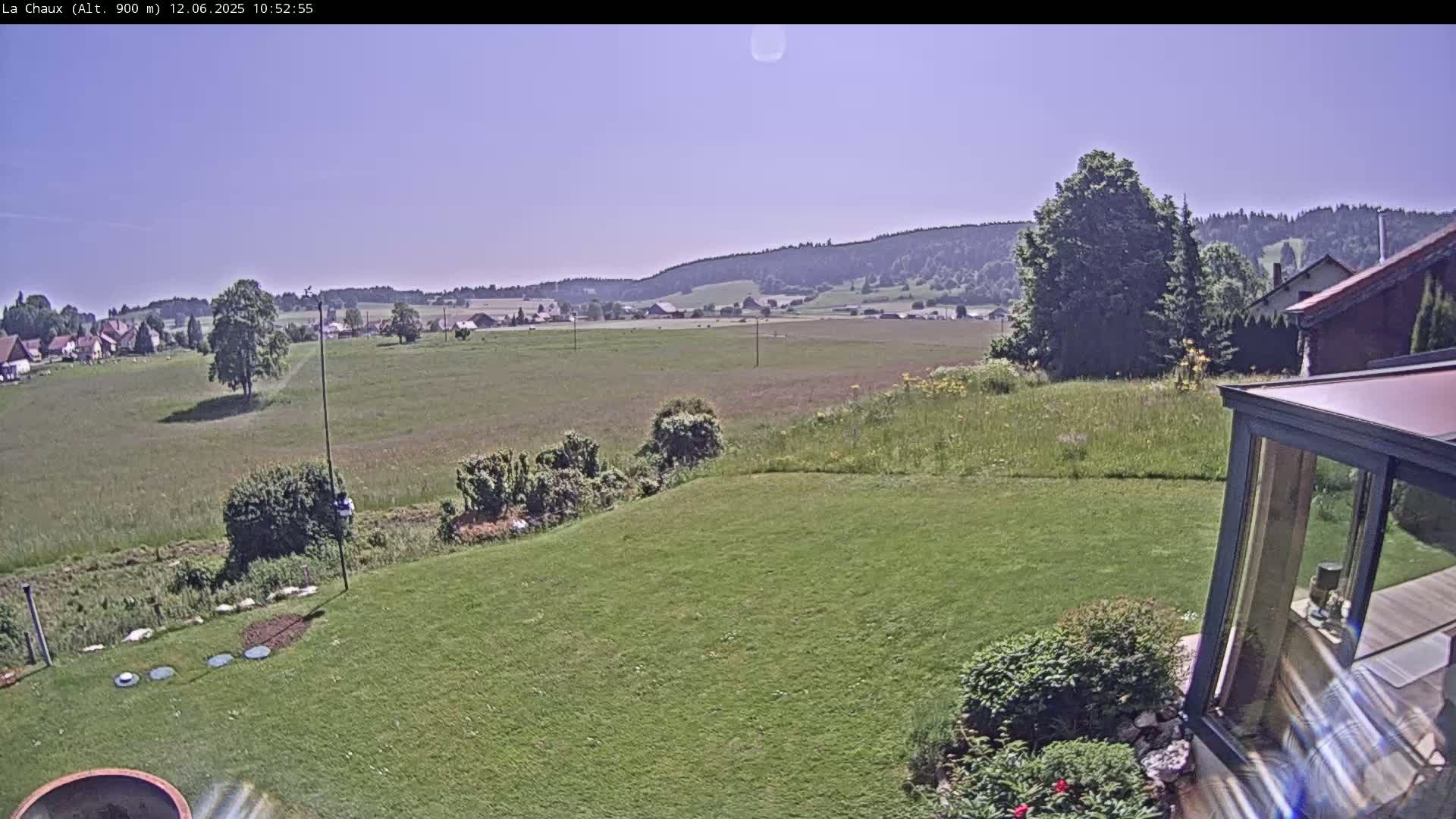 A sunny view from a backyard shows a grassy lawn sloping down to a field, beyond which lies a village nestled against rolling hills under a clear blue sky.