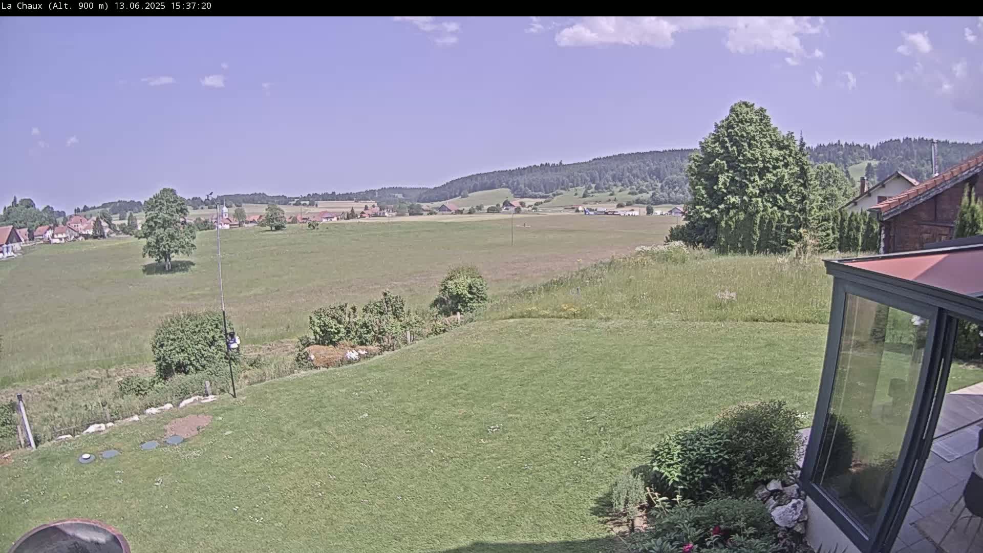A grassy yard with a modern glass enclosure overlooks a sprawling green field and a distant village under a mostly sunny sky.