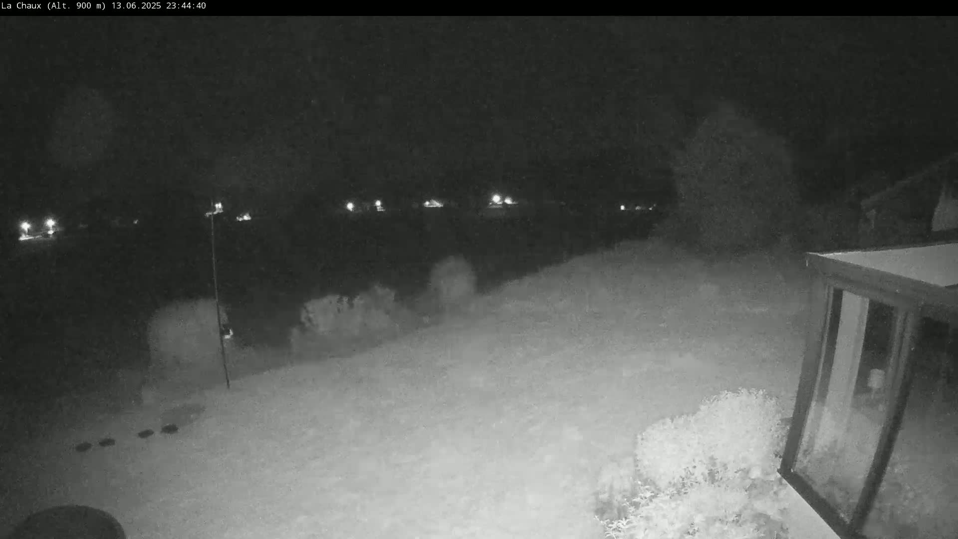 A snow-covered nighttime scene shows a backyard with some shrubs and a building, with distant lights visible in the background.