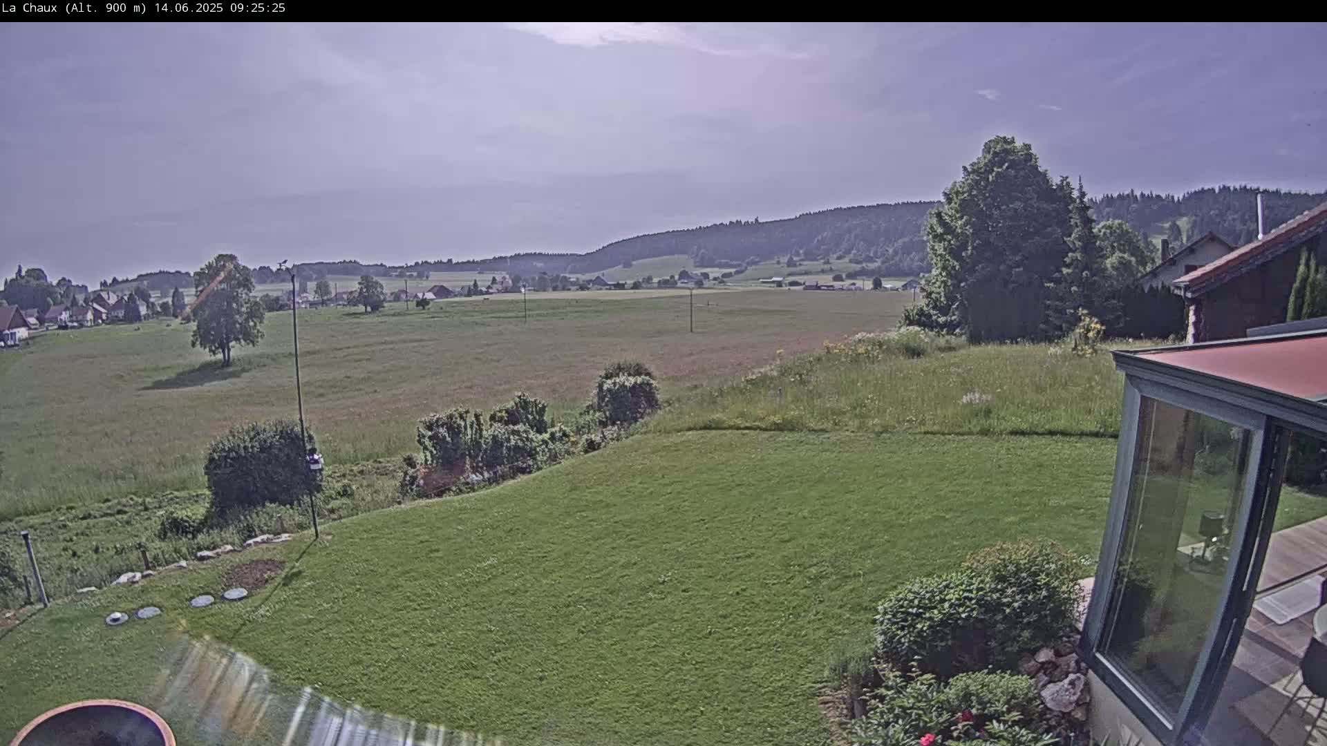 A mostly sunny view of a grassy yard, a house with a glass sunroom, and a distant village nestled in a valley surrounded by hills.