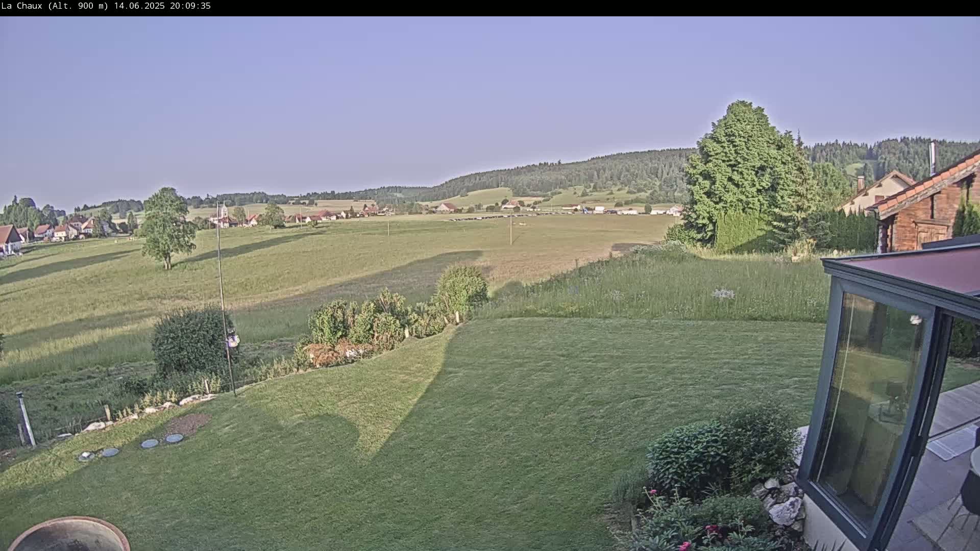 A grassy yard slopes down to a flat field and village, under a clear, light-blue sky.