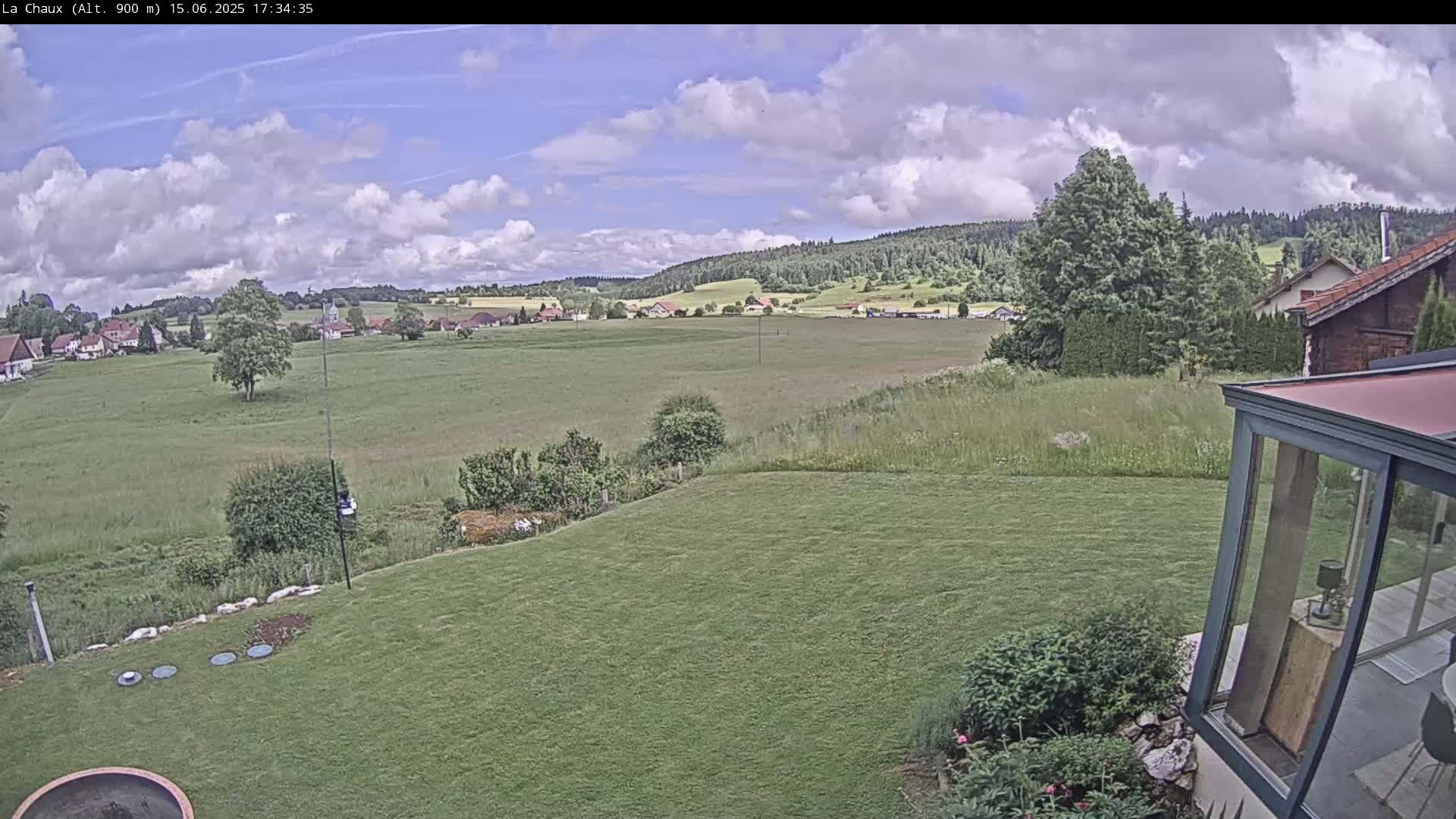 A partly cloudy day reveals a sprawling green field, a distant village nestled among rolling hills and trees, and a section of a well-maintained lawn with a modern glass structure in the foreground.