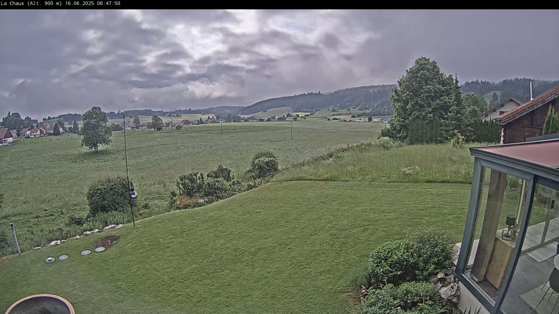 A view from a backyard shows a grassy lawn sloping down to a verdant field, beyond which sits a village nestled in a valley under a cloudy sky.