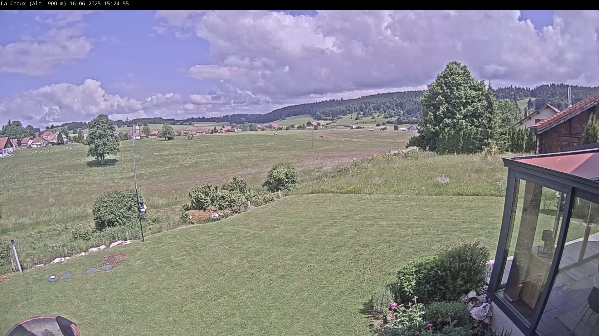 A partly cloudy day reveals a verdant landscape featuring a sprawling green lawn, a distant village nestled amidst fields, and a backdrop of rolling hills covered in forest.