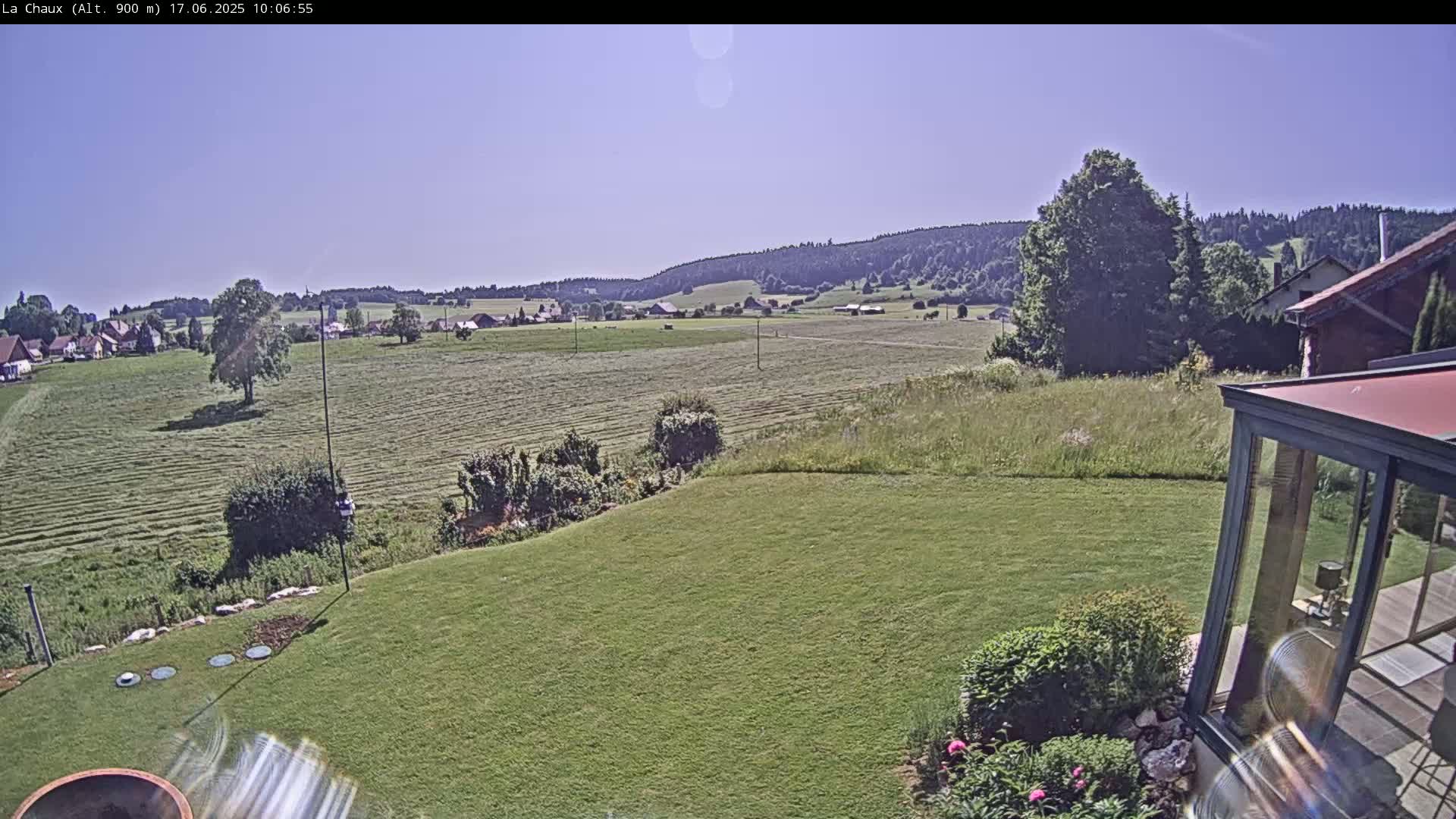 A sprawling green lawn slopes down to a mown field under a clear, sunny sky, with a village and distant hills visible in the background.