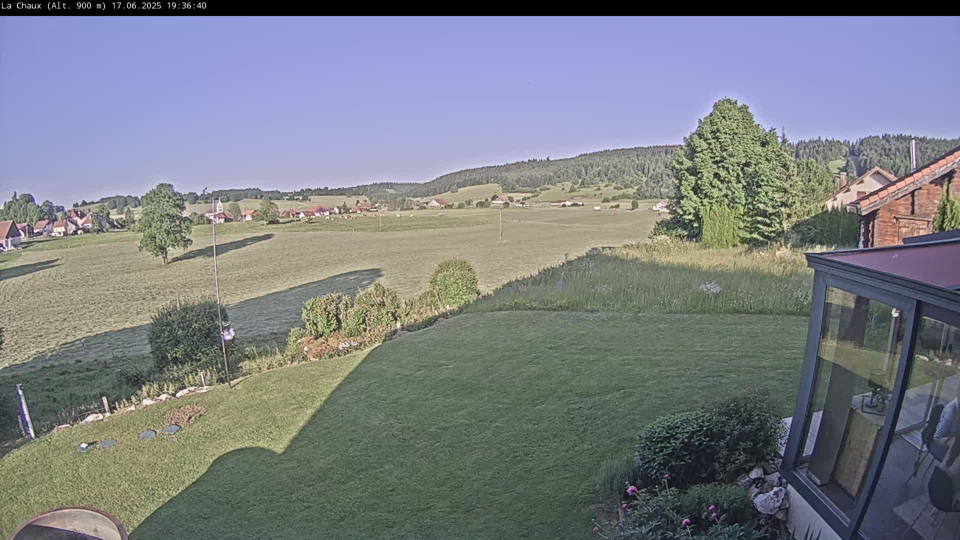 A clear, sunny day reveals a verdant landscape of fields and a village in the distance, viewed from a garden with a glass conservatory.