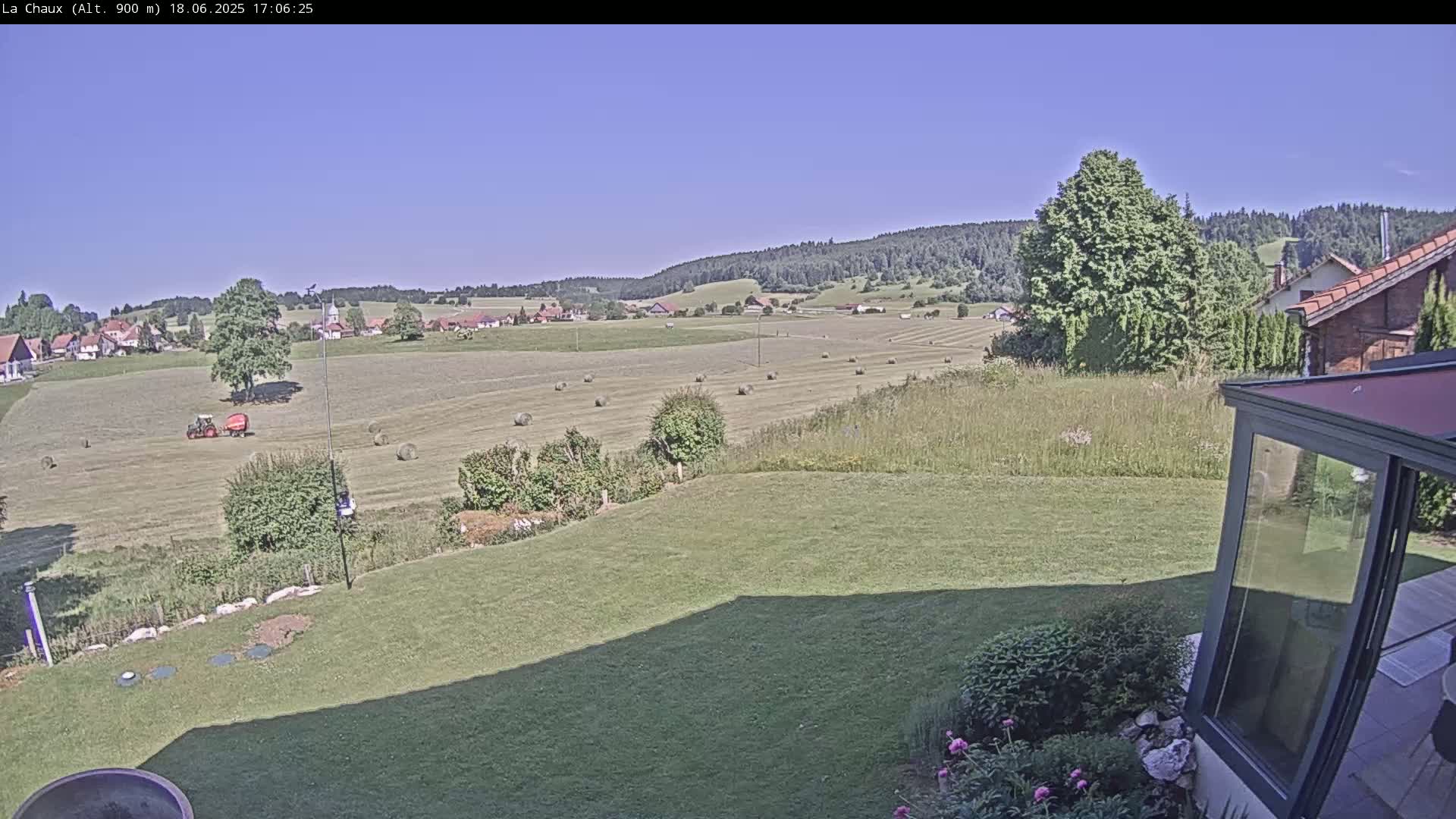 A sunny view from a grassy backyard shows a field of hay bales, a tractor, a small village, and a forest-covered hillside in the distance.