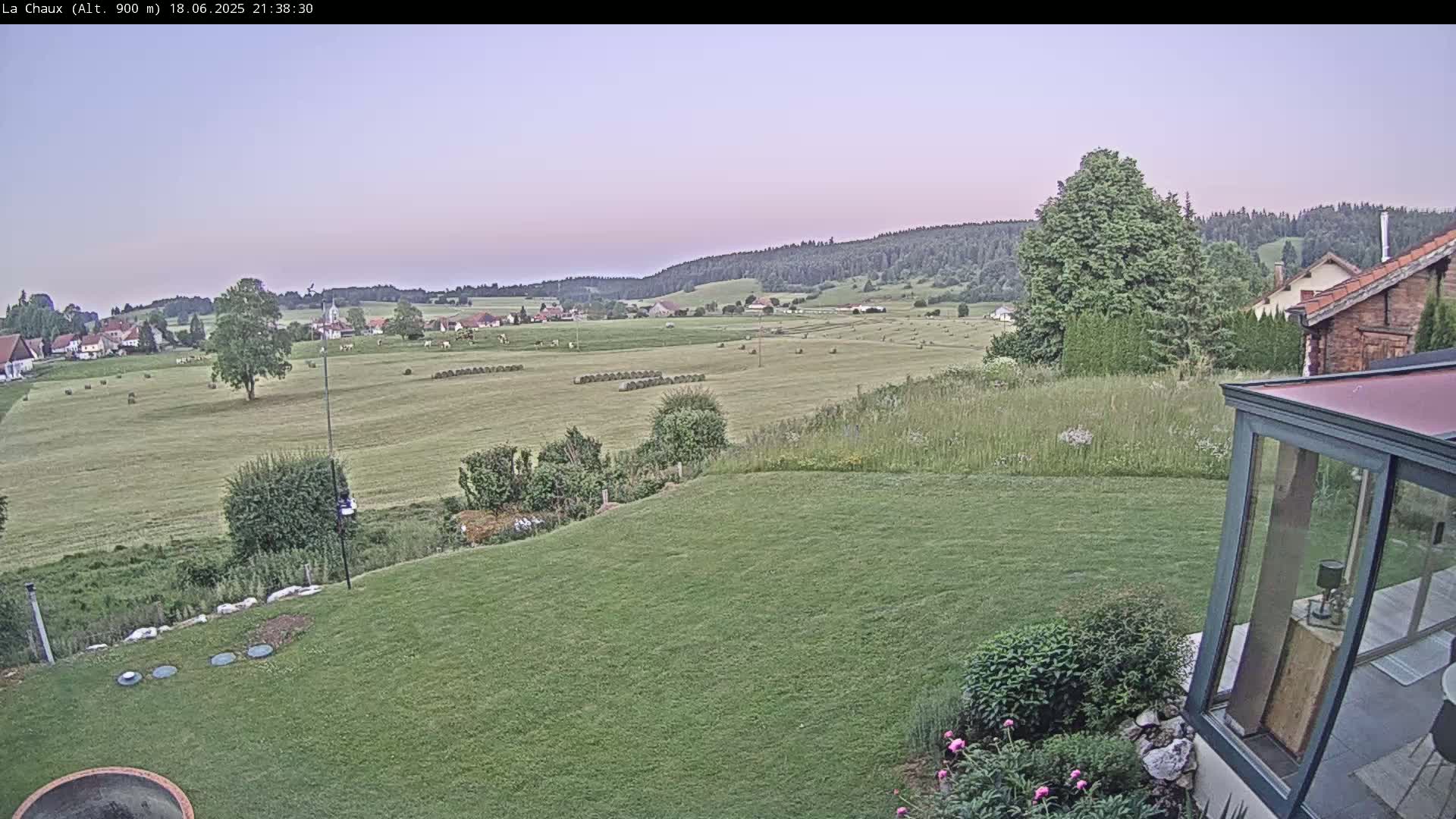 A sprawling green lawn slopes down to a field dotted with hay bales, under a twilight sky, with a village and forested hills visible in the distance.