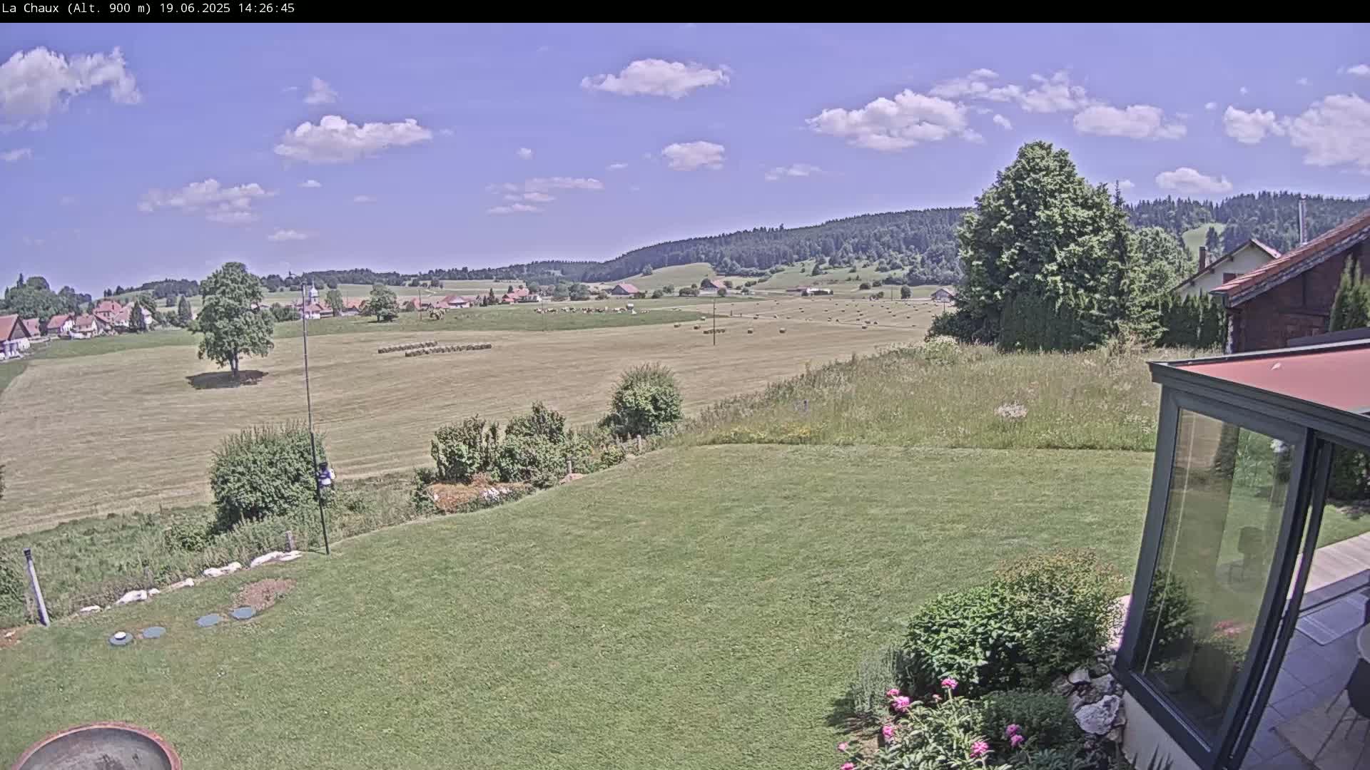 A sunny day reveals a verdant landscape of hay bales in a field, a village nestled in the distance, and a grassy yard with a modern glass enclosure in the foreground.