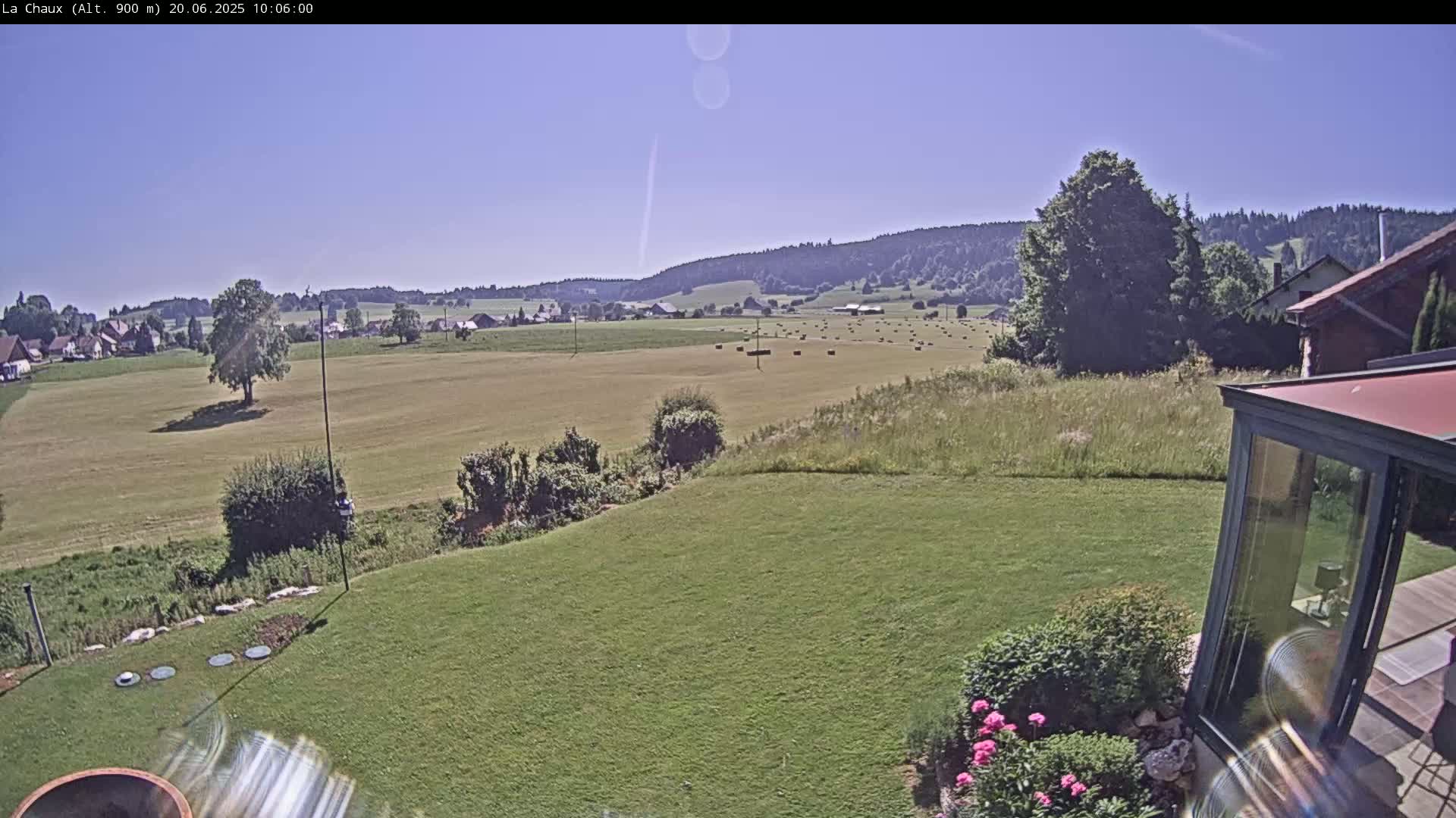A sunny view from a backyard shows a grassy field with hay bales in the distance, beyond which are rolling hills and a village under a clear sky.
