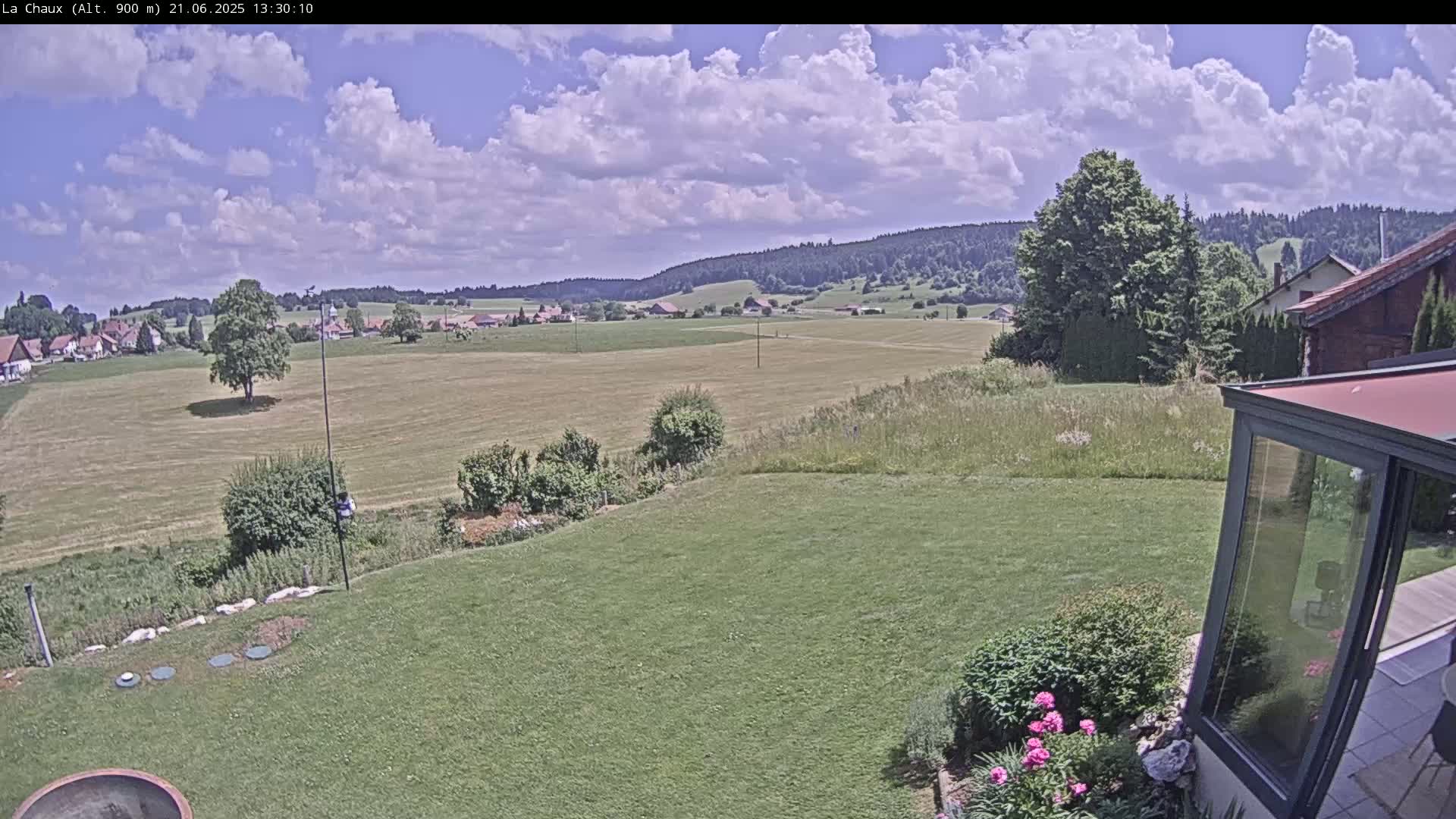 A partly cloudy day reveals a sprawling green field, a distant village nestled in a valley, and a verdant lawn bordered by a modern glass enclosure.