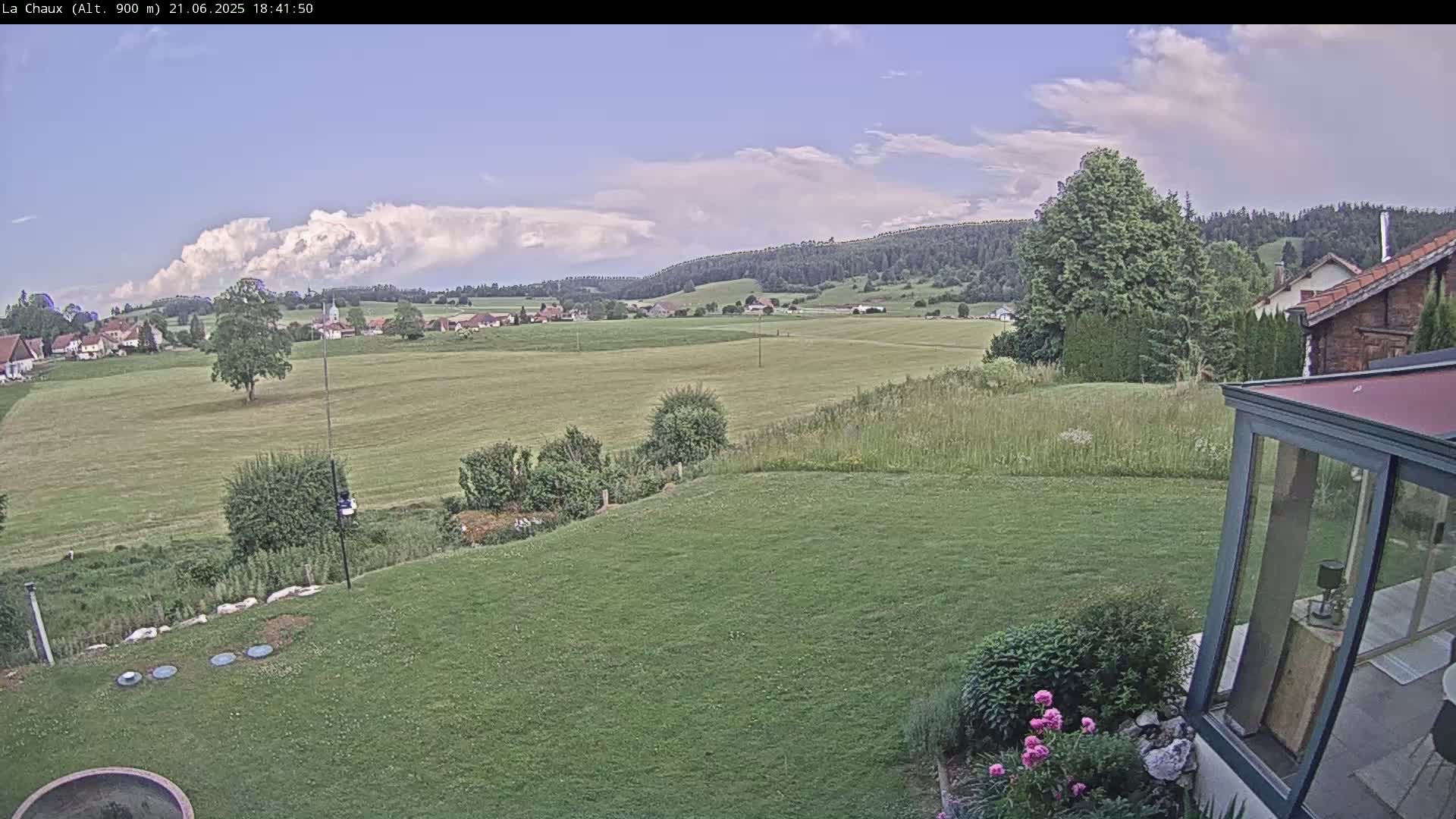A partly cloudy sky overlooks a grassy field, a small village, and rolling hills, as seen from a backyard with a modern glass enclosure.