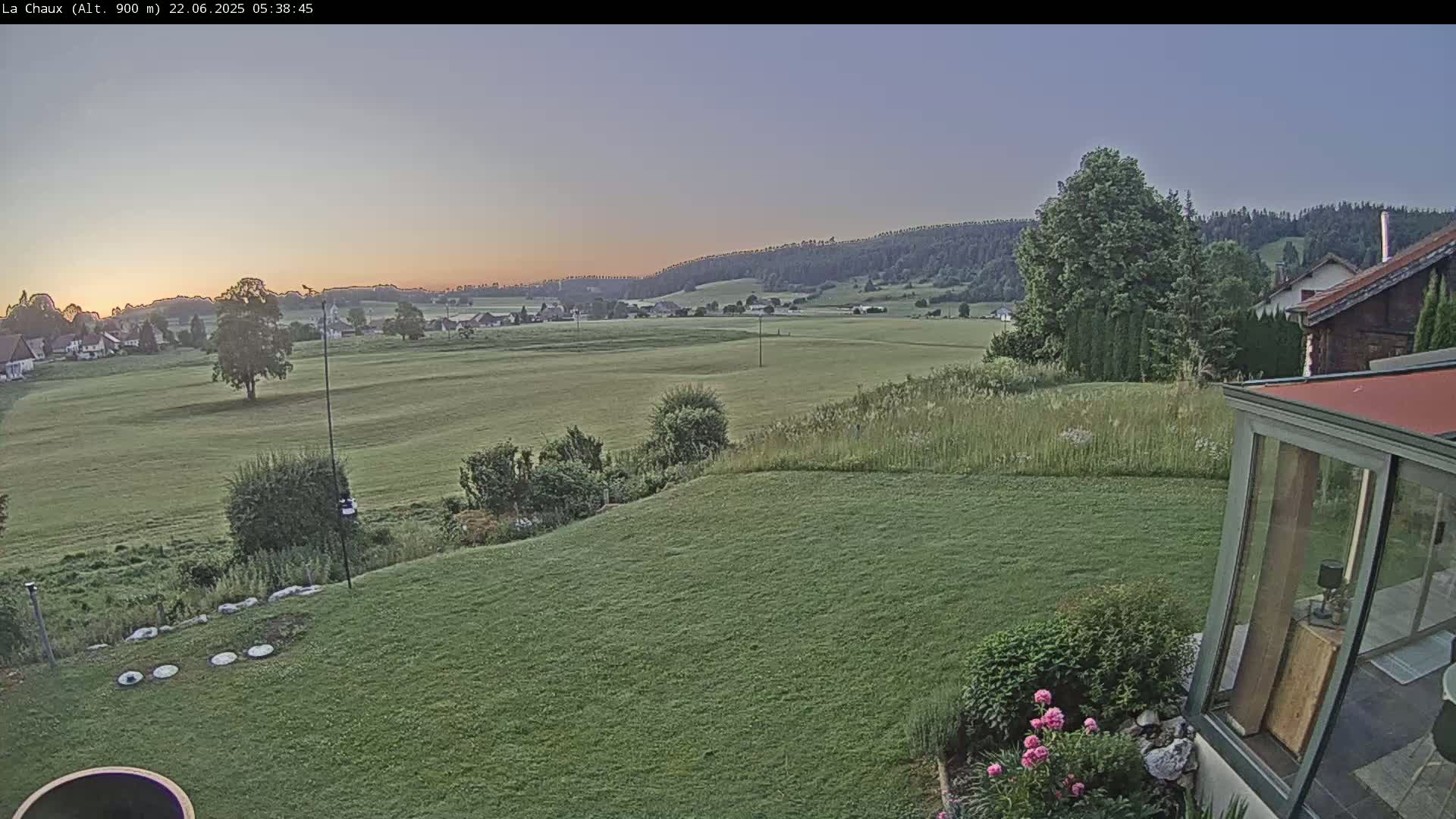 A sunrise illuminates a grassy yard and distant rolling hills under a clear sky, with a portion of a house and garden visible in the foreground.