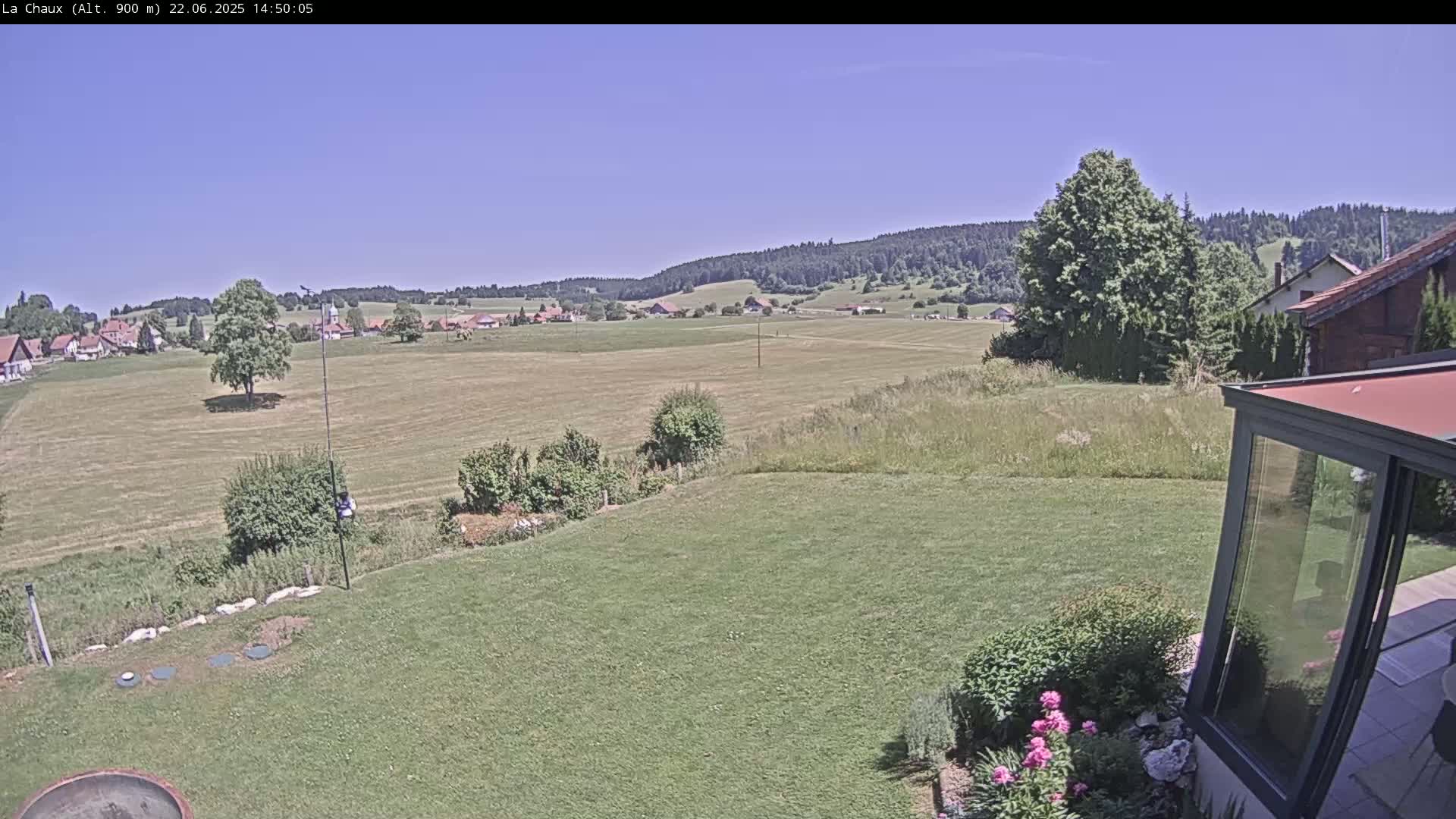A sunny view of a grassy yard and a distant village nestled in a valley, backed by a line of dark green hills.