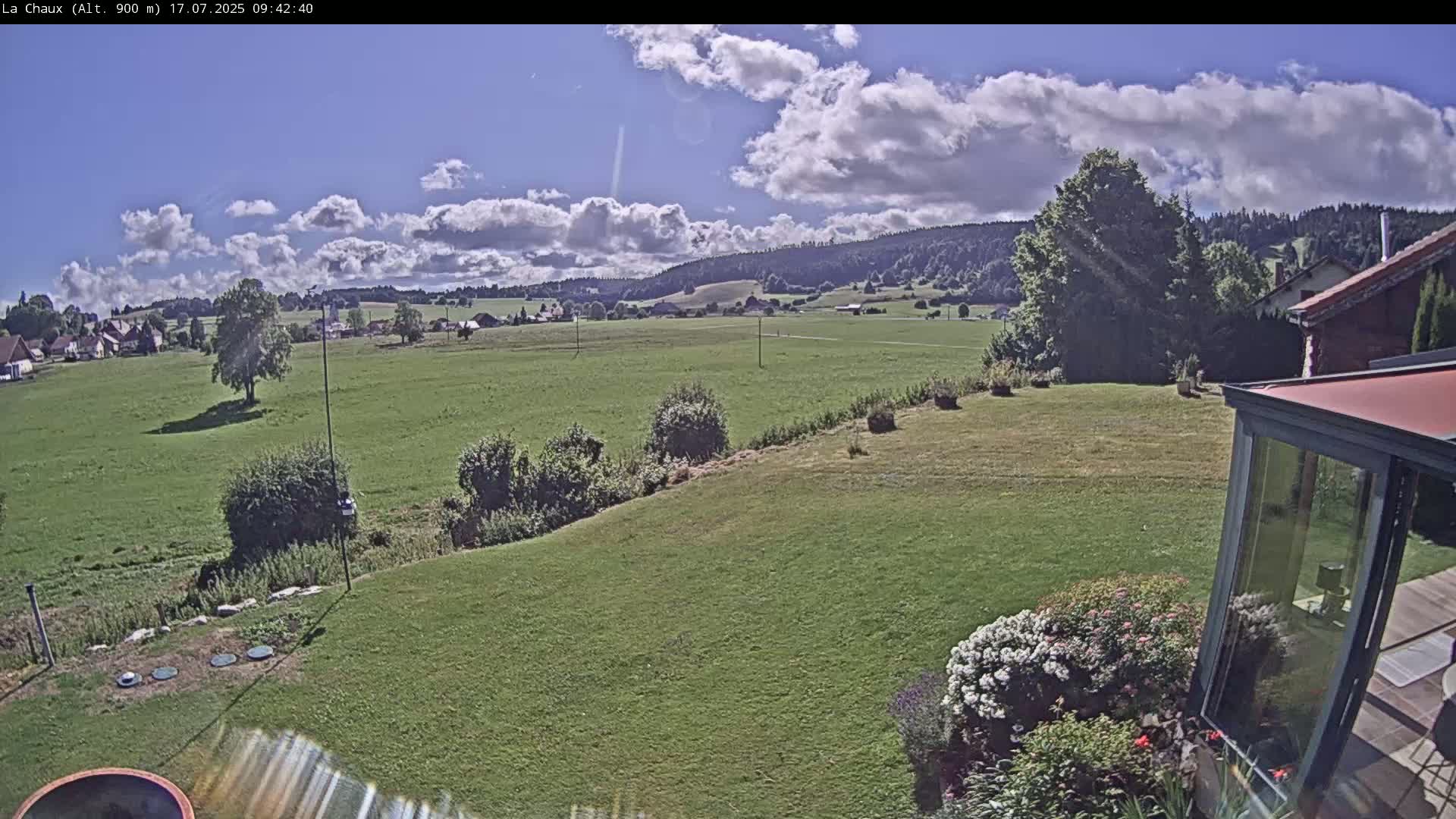 A partly cloudy sunny day reveals a sprawling green field, a small village in the distance, and a portion of a house with a sunroom in the foreground.