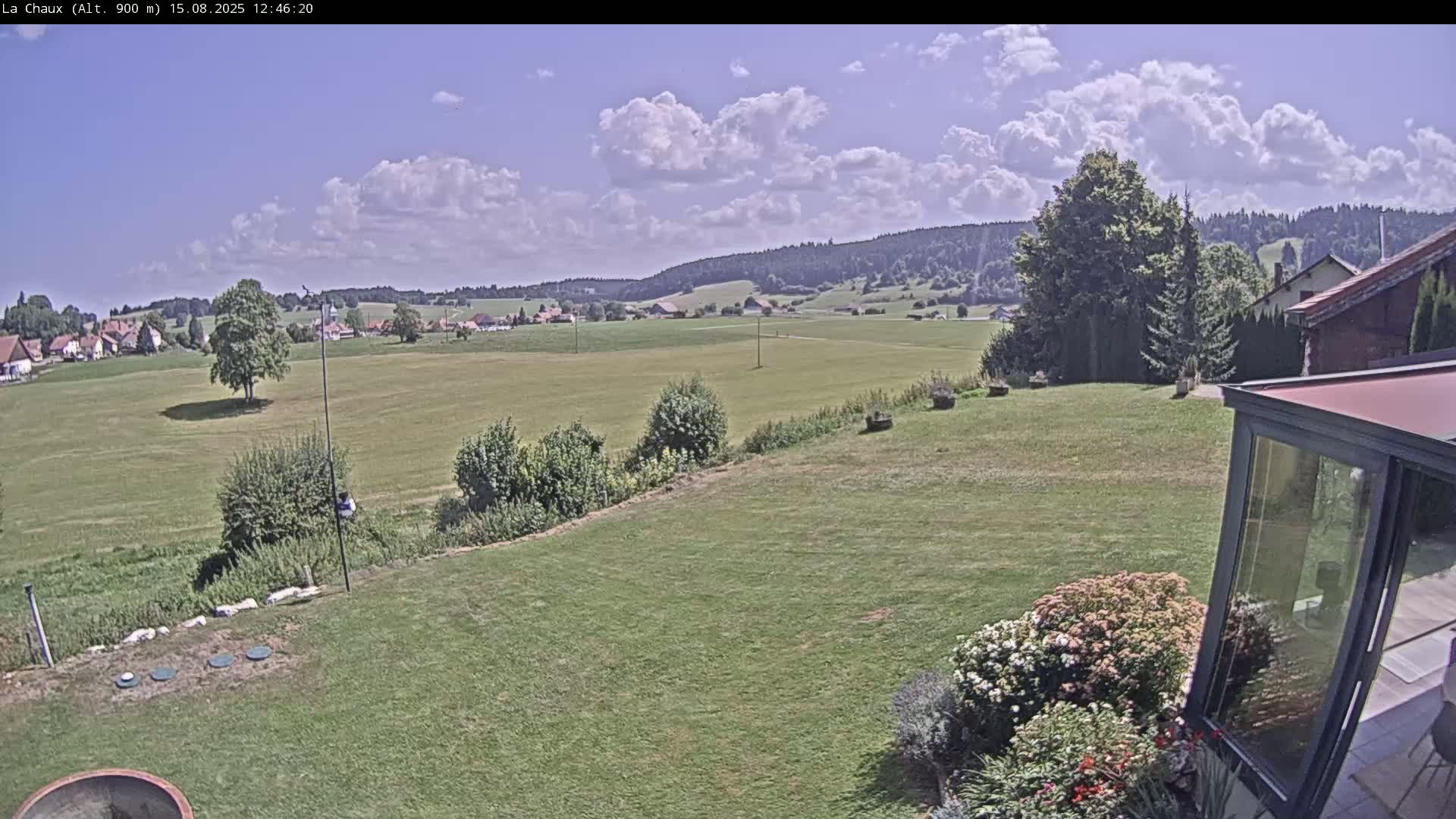 A partly cloudy sunny day reveals a sprawling green field, a small village in the distance, and a portion of a house with a sunroom in the foreground.