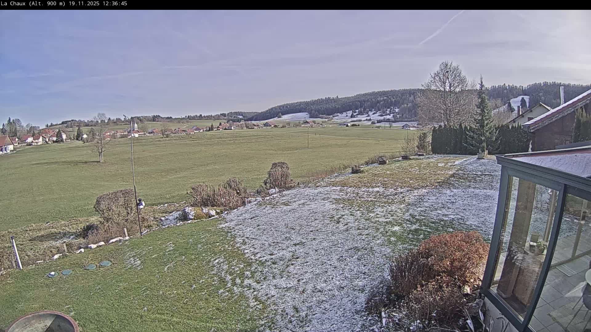 A partly cloudy sunny day reveals a sprawling green field, a small village in the distance, and a portion of a house with a sunroom in the foreground.